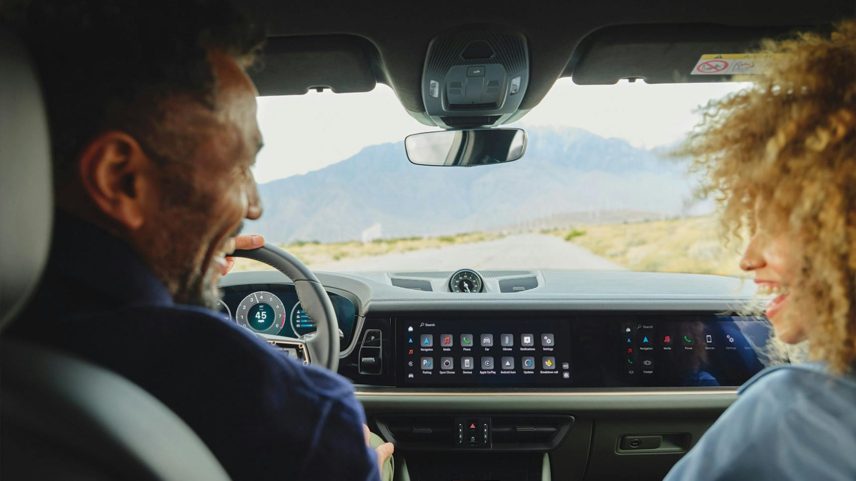 Interior view of a Porsche car showing a modern dashboard with touchscreen displays and navigation features, while two passengers enjoy a scenic drive on a straight road leading toward distant mountains.
