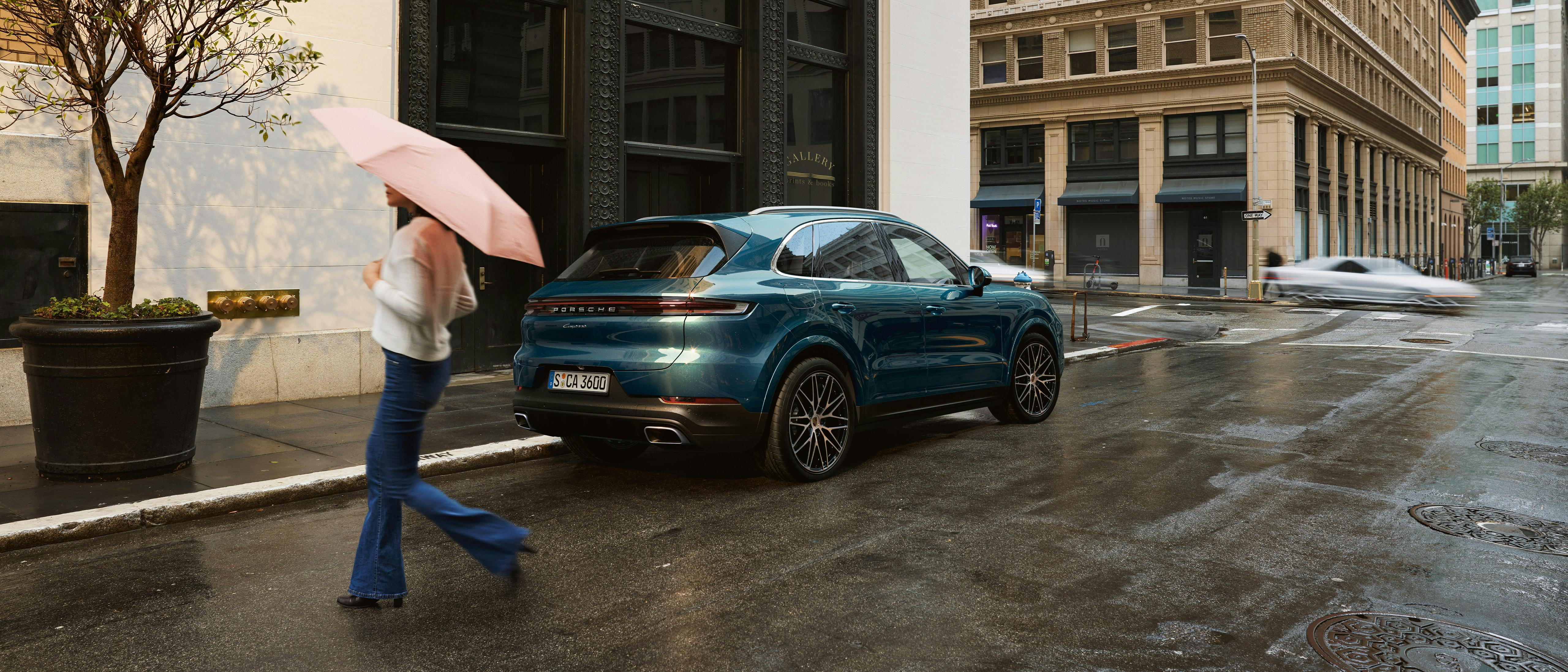 Une Porsche Cayenne bleue est garée sur le côté mouillé par la pluie. Une femme avec un parapluie traverse la rue.