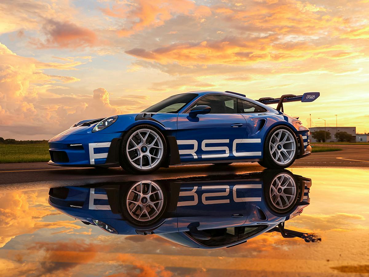 Porsche 911 GT3 RS Weissach Package in blue with white motorsport graphics, photographed at sunset with dramatic sky and reflection on wet pavement for a dynamic performance shot.