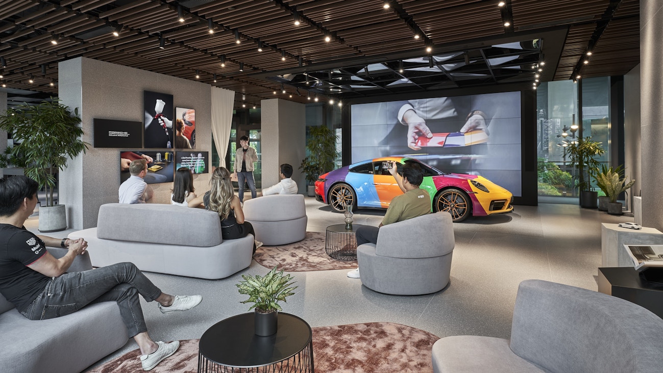 Image 6: Exhibit area at Porsche Studio Singapore with guests listening to a presentation, a colourful Porsche 911 Carrera GTS on display.
