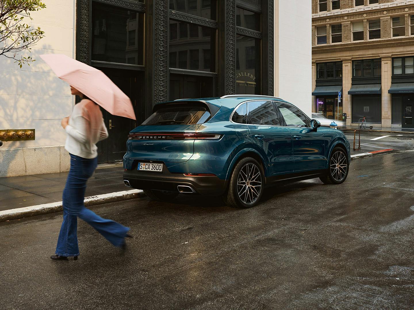 Ein blauer Porsche Cayenne ist an der regennassen Seite geparkt. Eine Frau mit Regenschirm kreuzt die Straße.