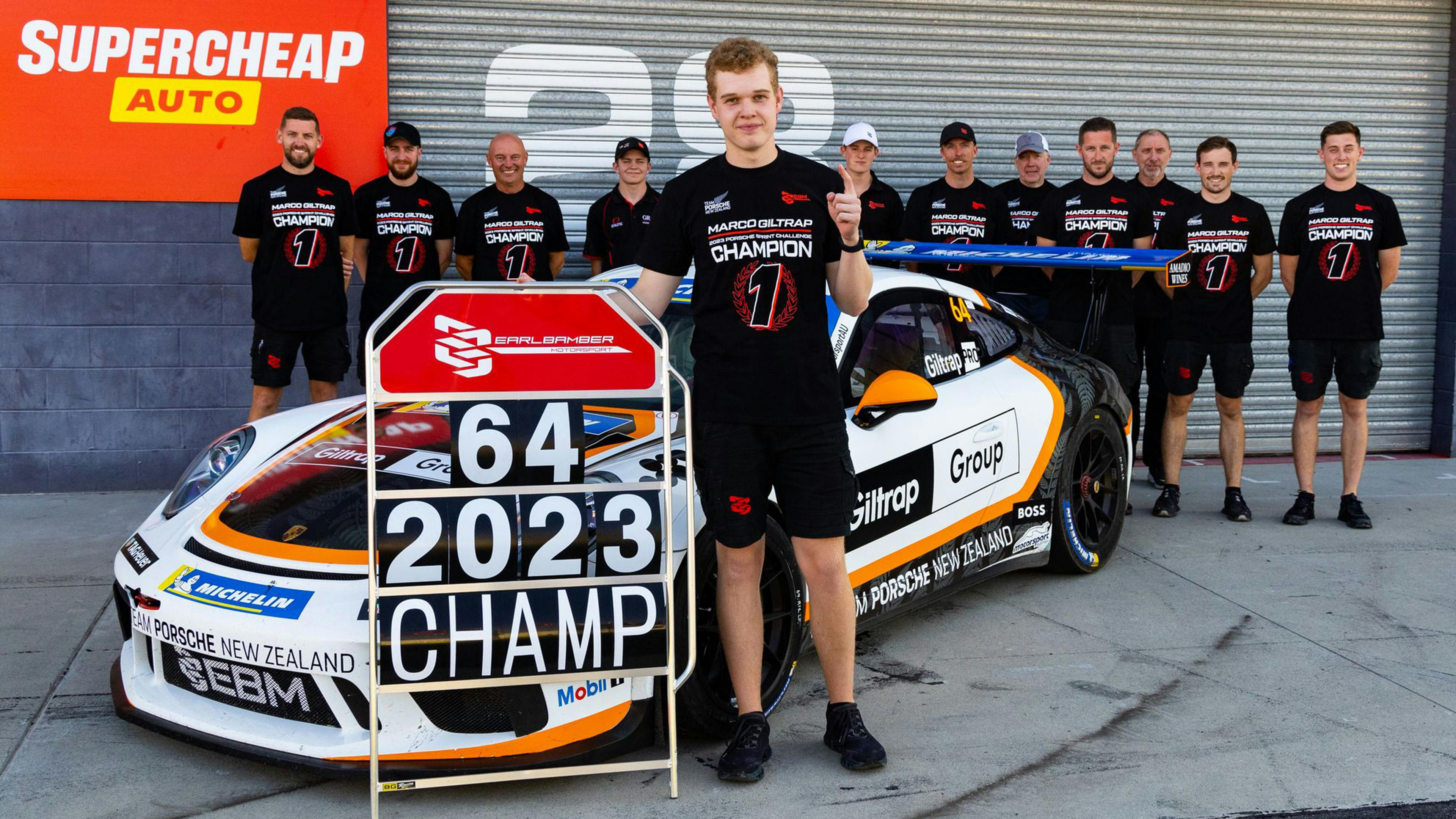 Marco Giltrap stands in front of a Porsche holding his starting and year number as champion, with his team behind him.