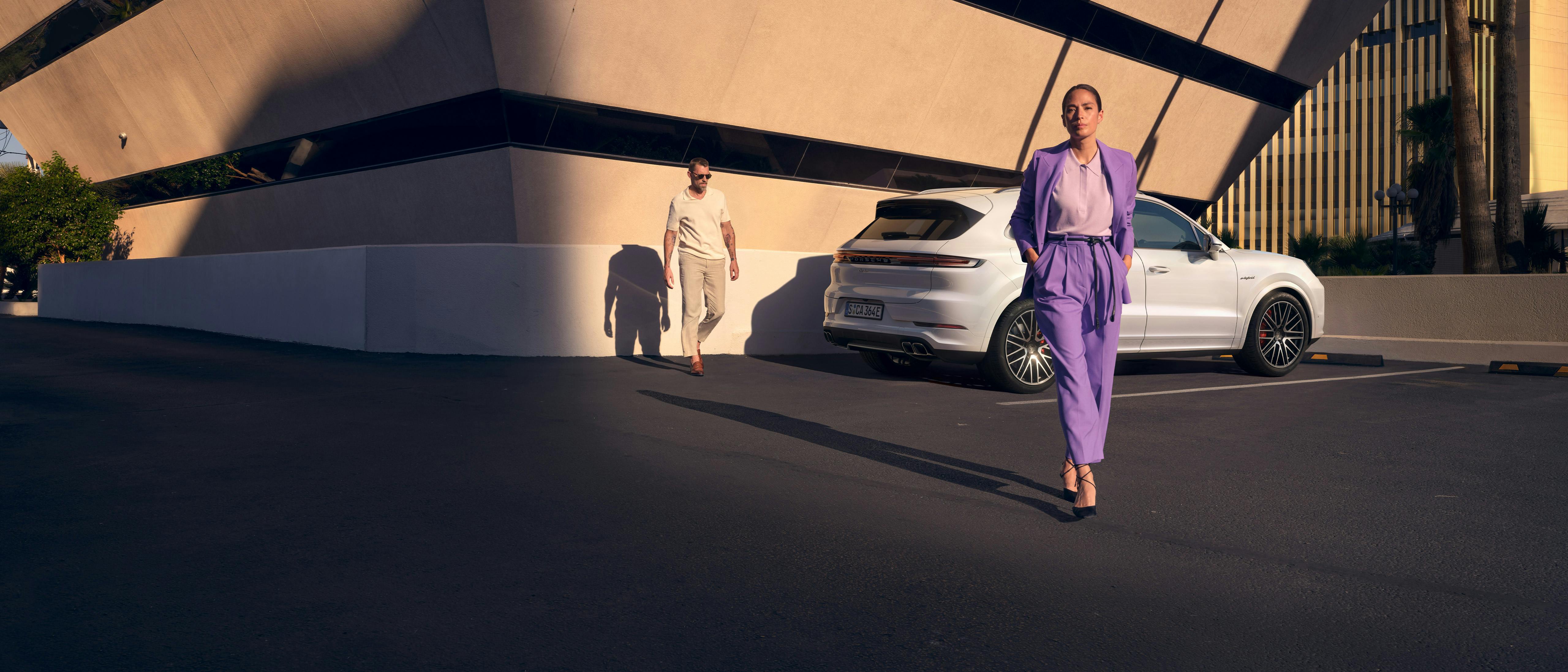 Un hombre y una mujer posan delante de un moderno edificio junto a un Cayenne Turbo E-Hybrid blanco.