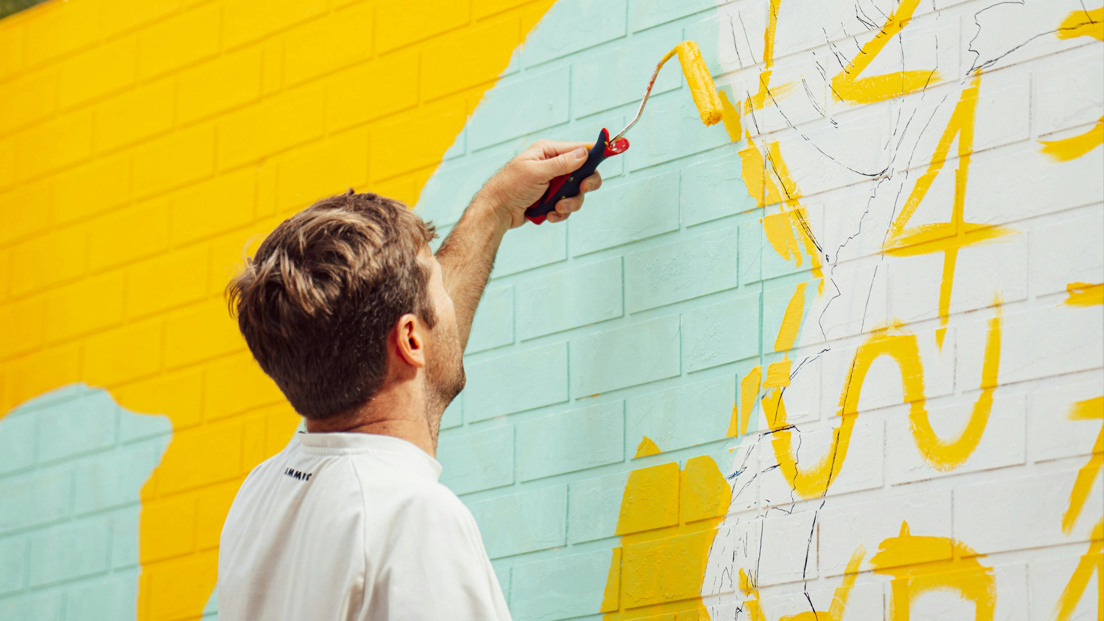 An artist with his back to the camera uses a paint roller to apply bright yellow paint to a brick wall. The wall features a large-scale mural in progress, with sections of light blue and yellow paint overlapping.​