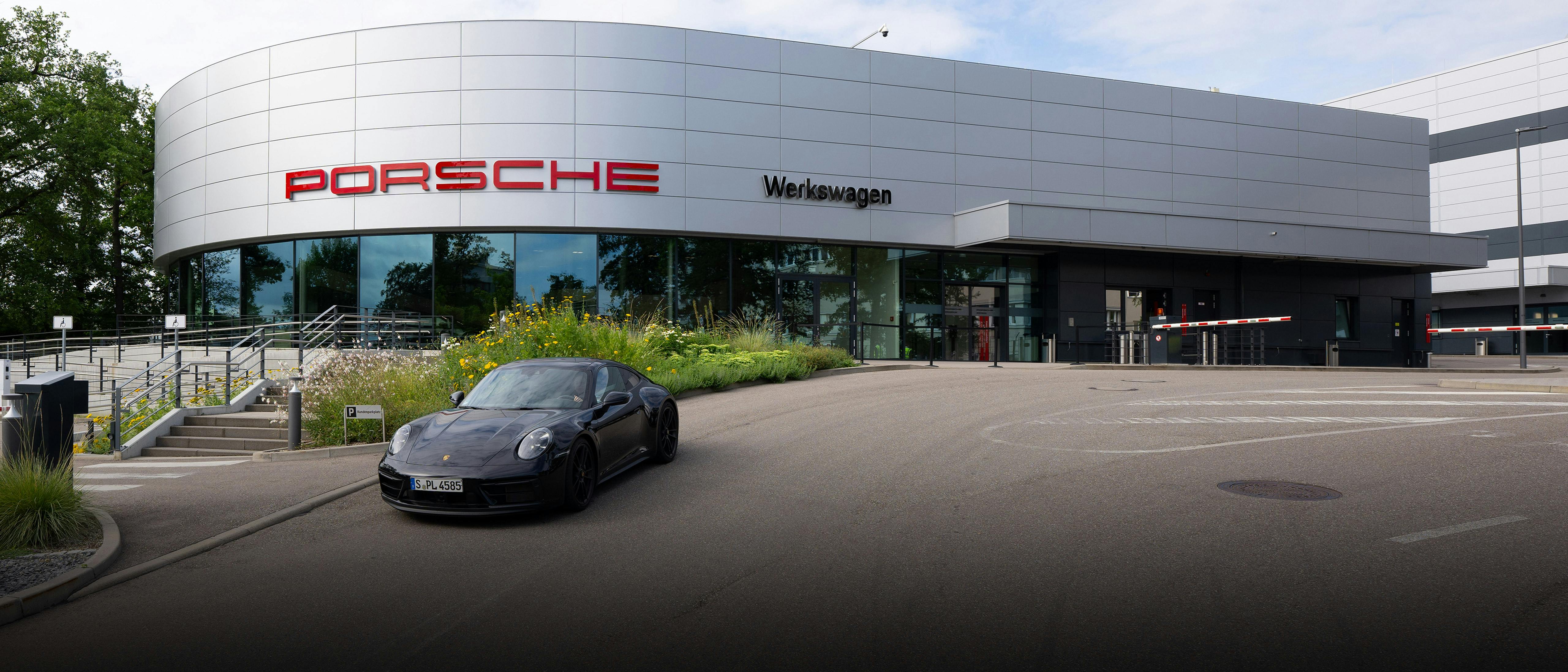 Schwarzer Porsche 911 vor modernem Werkswagen-Gebäude mit rotem Schriftzug und Glasfassade.