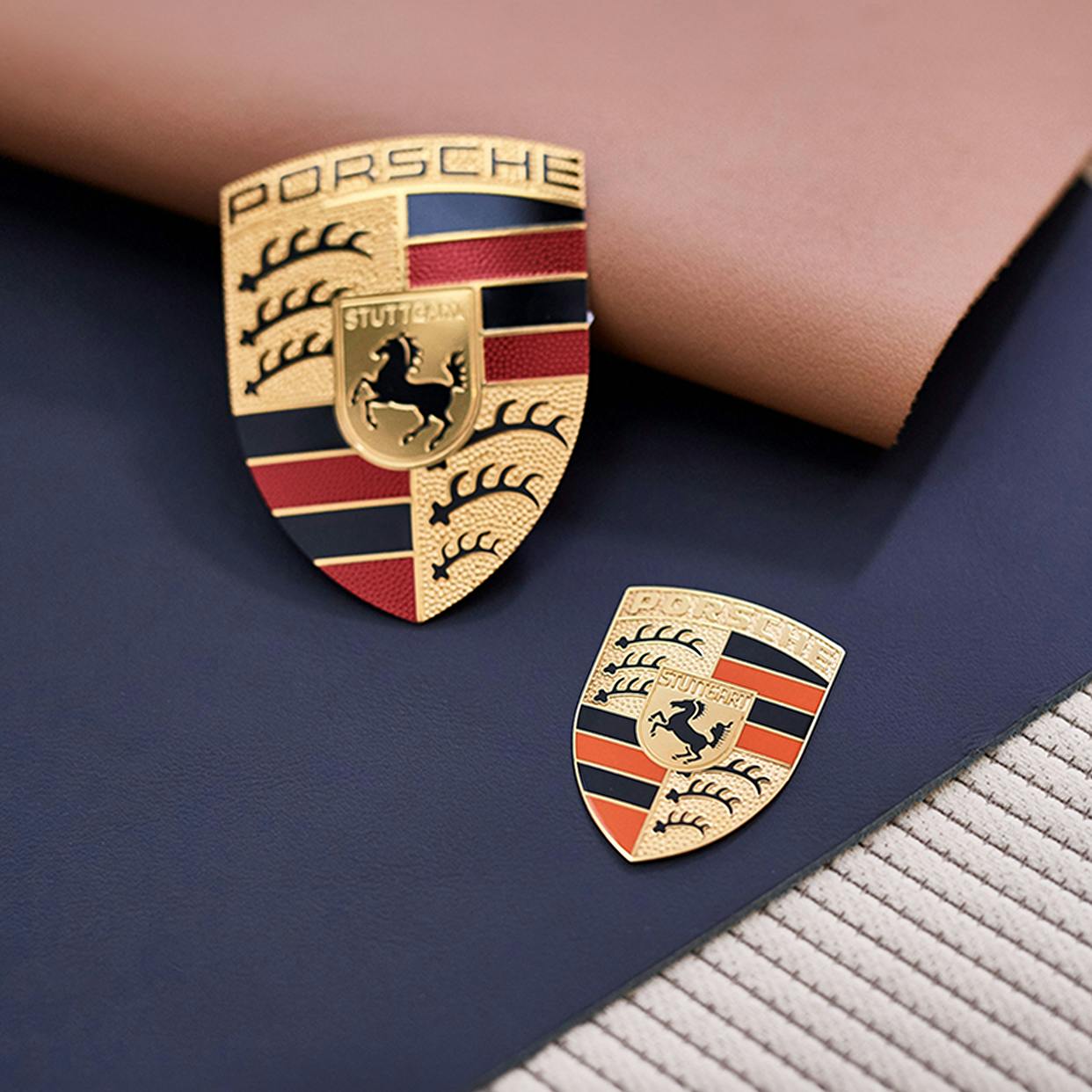 2 Porsche emblemen in verschillende maten liggen op stoffen en lederen patronen.