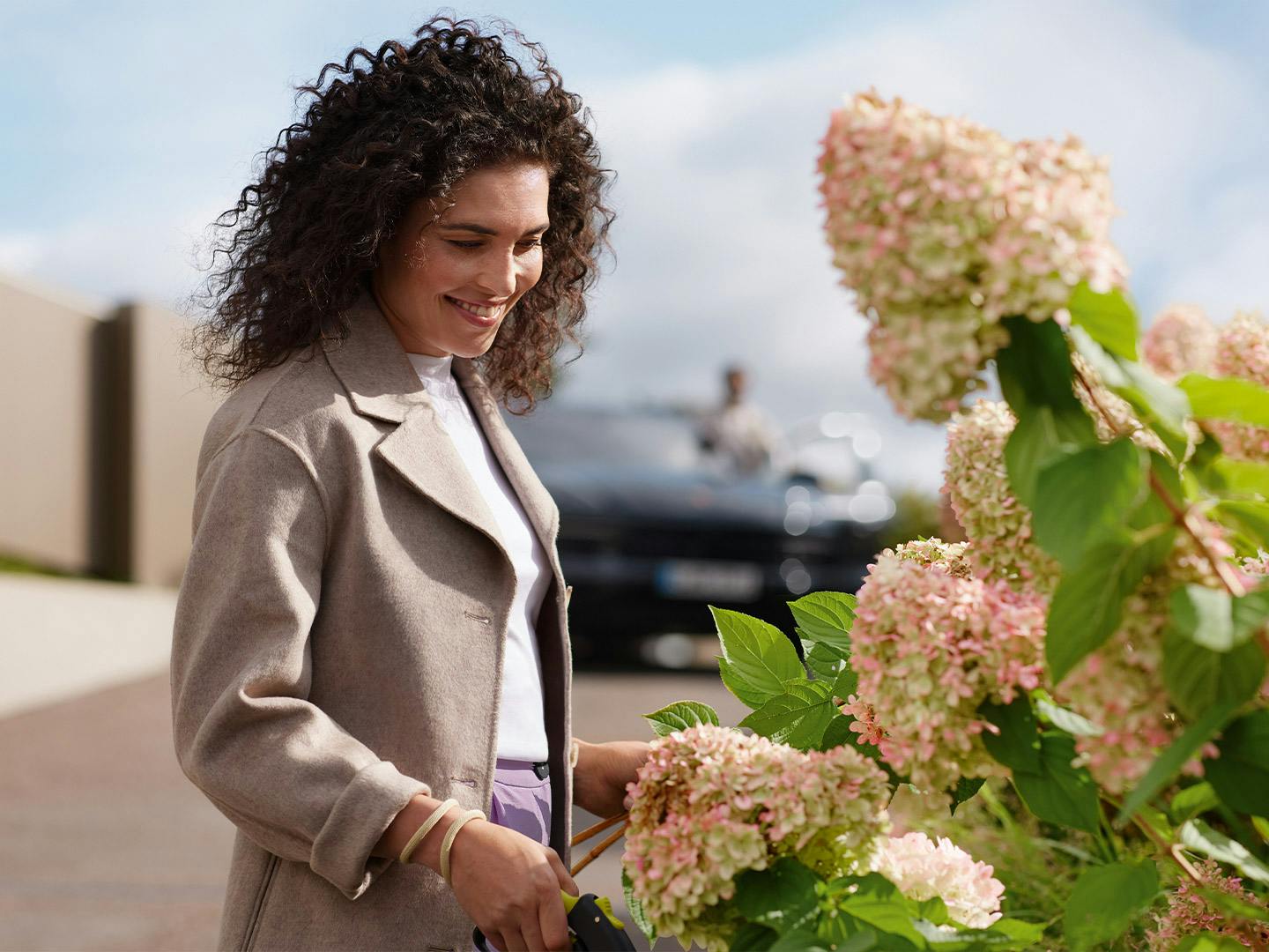 Une femme regarde des hortensias en fleurs. En arrière-plan, on aperçoit vaguement une Porsche.