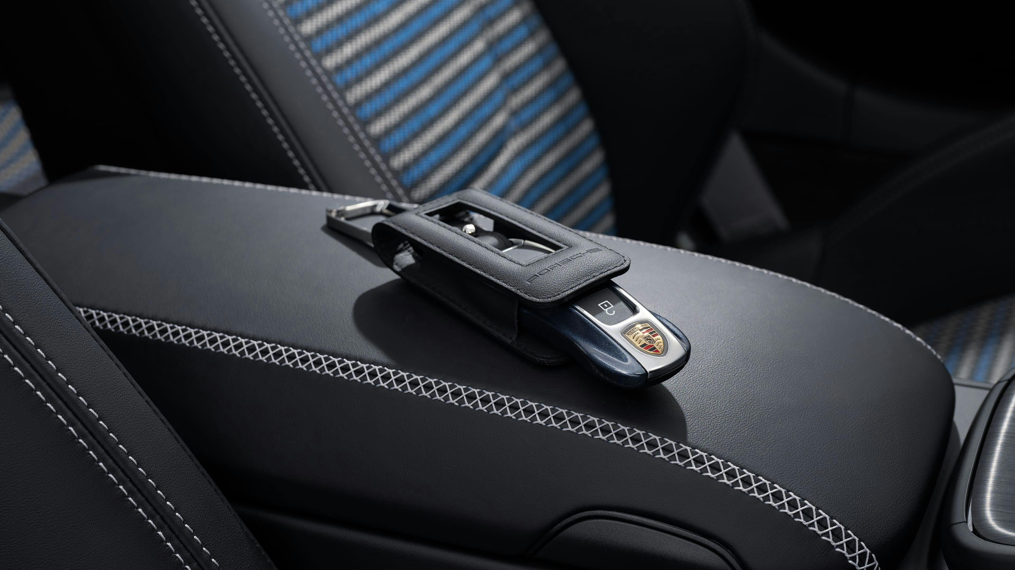 Porsche key in a leather holder resting on a black centre armrest, with blue-patterned sports seats in the background.