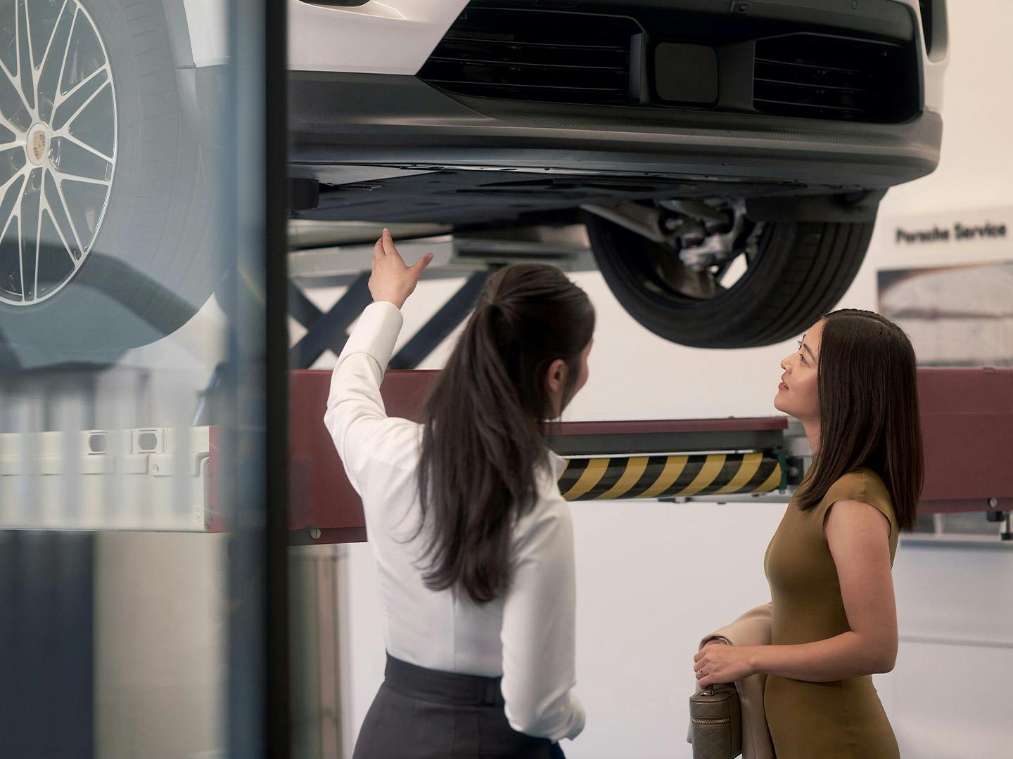 Porsche service advisor having a conversation with a customer while inspecting their Porsche on a vehicle lift in a workshop.