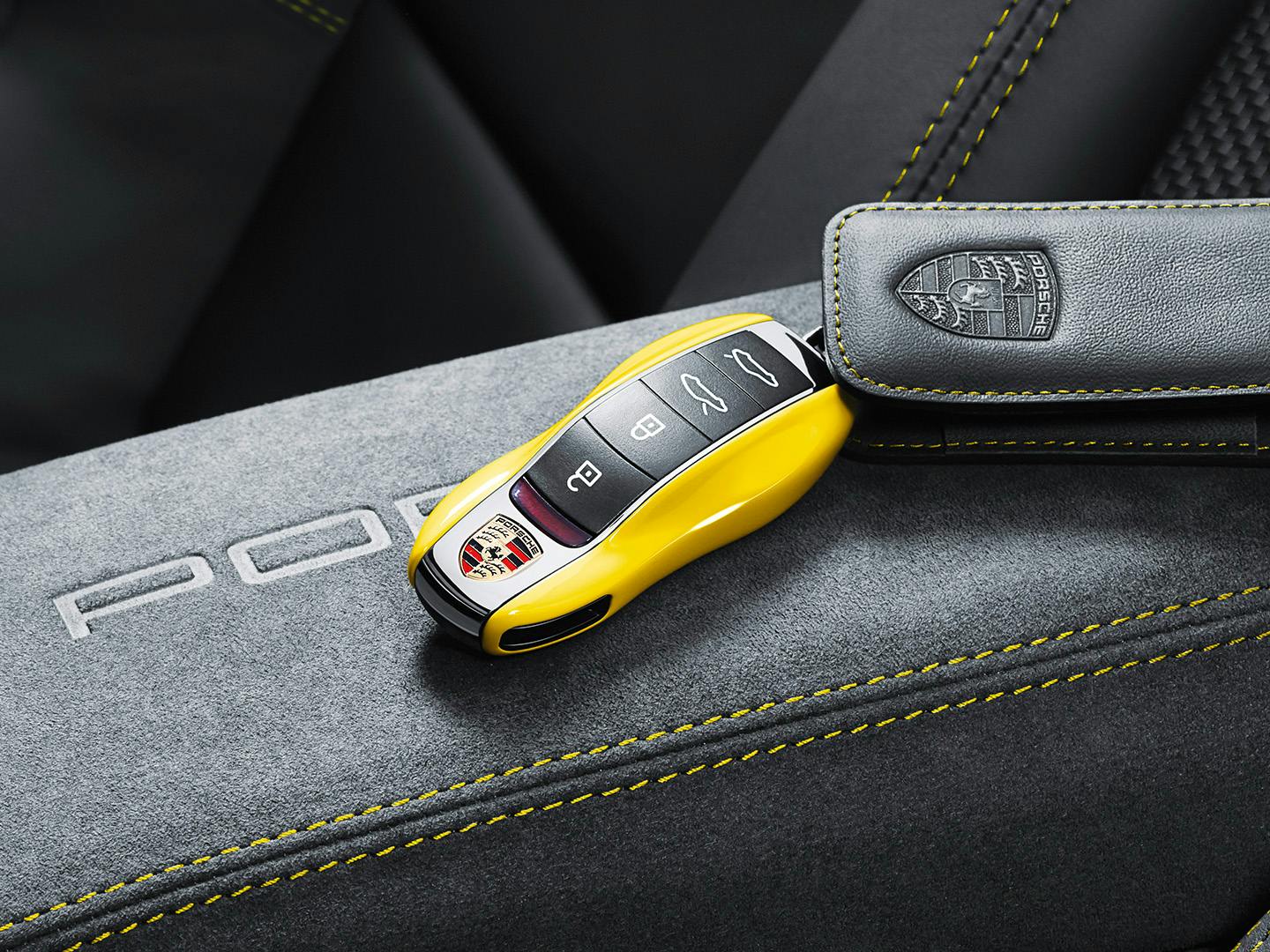 Yellow Porsche car key fob with logo and buttons placed on a gray interior surface with yellow stitching, accompanied by a leather key holder.