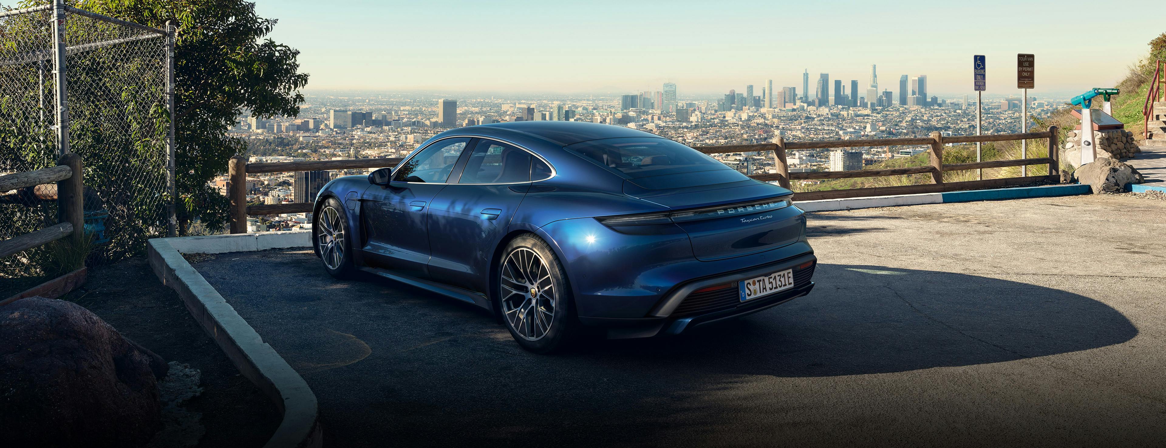 A man is leaning on a metallic blue Porsche Taycan Turbo, using Apple Maps EV Routing on his iPhone.