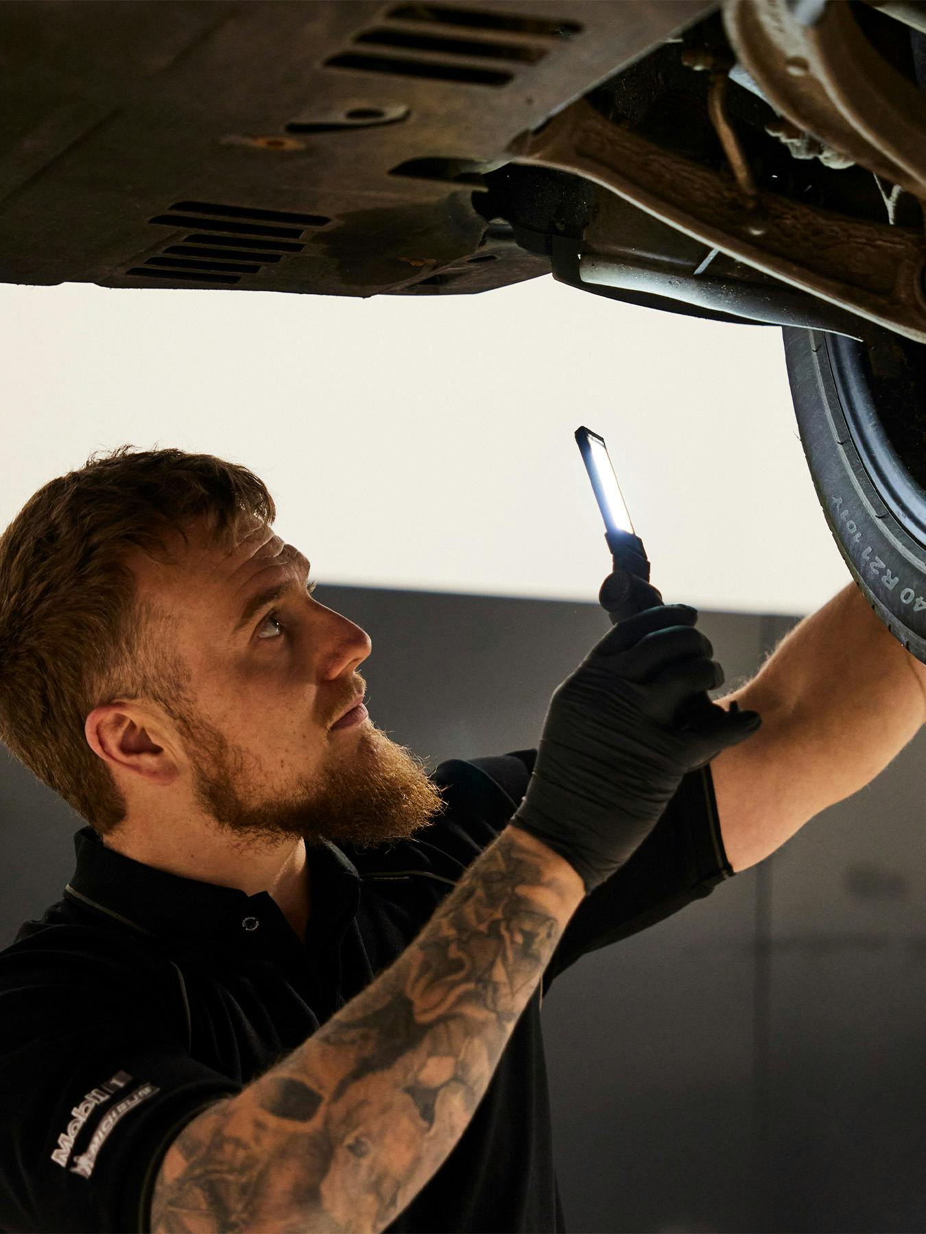 A mechanic with a beard and a tattooed arm stands beneath a vehicle on a lift. He is wearing black gloves and a black polo shirt, holding a slim LED work light to inspect the front suspension and wheel assembly. The focus is on his concentrated expression as he illuminates the mechanical components.