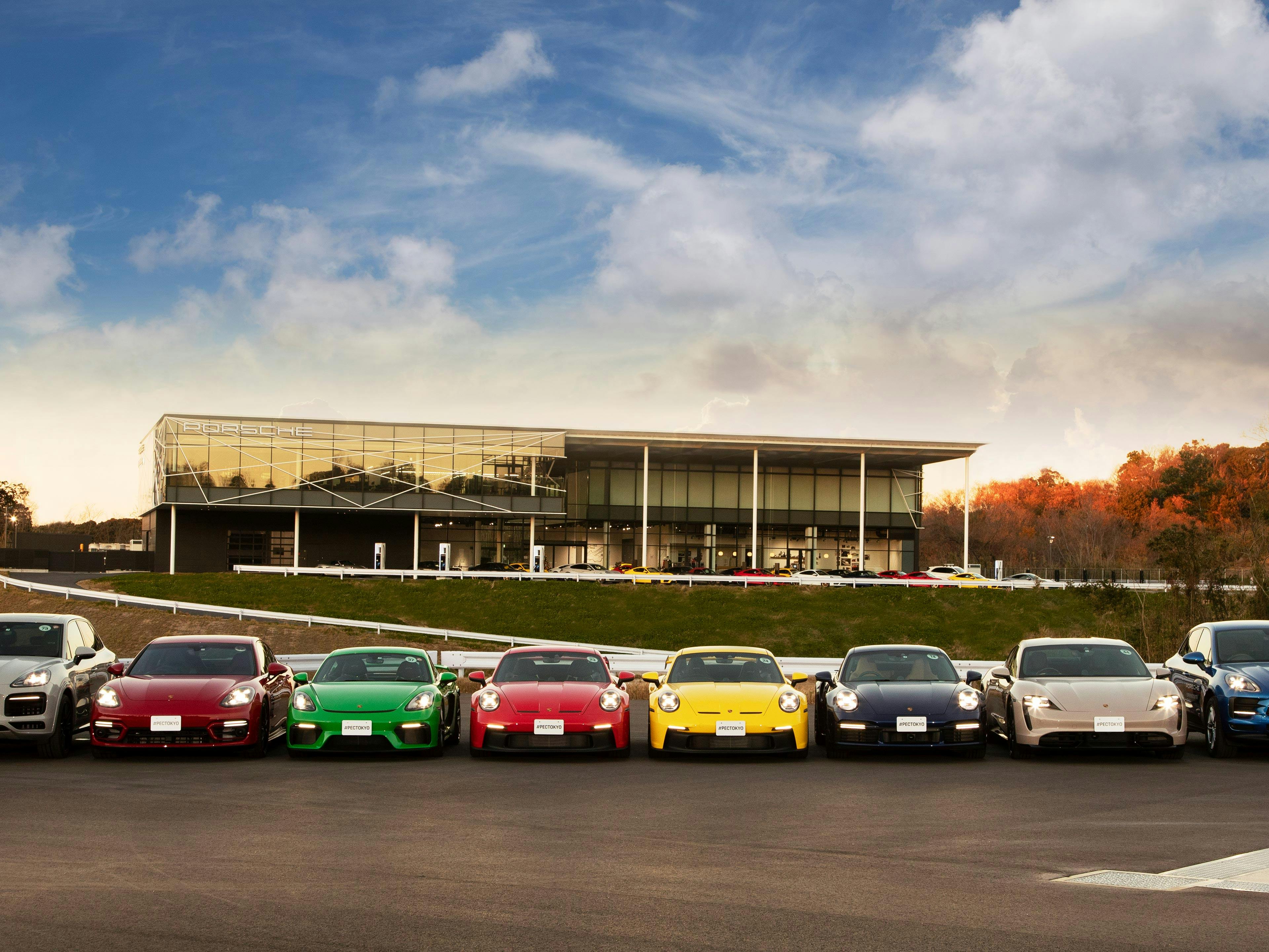 Ocho vehículos Porsche se encuentran uno al lado del otro, formando una fila sobre el asfalto; al fondo, el edificio del Porsche Experience Center de Tokio.