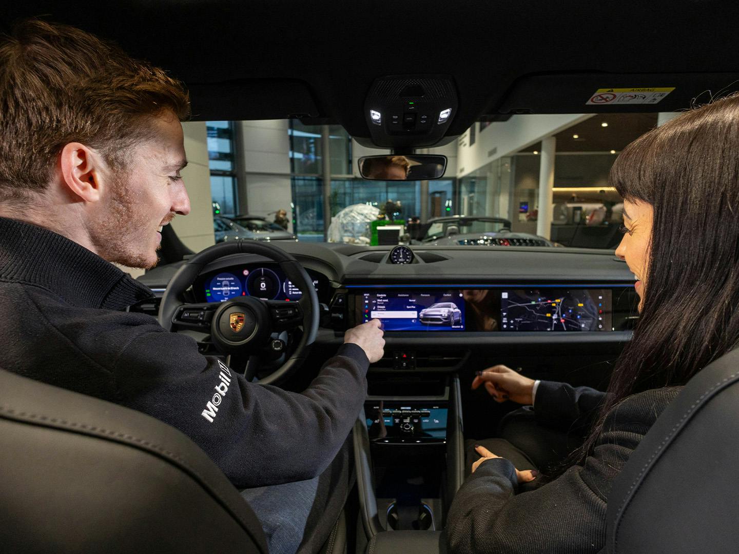 Un employé de Porsche, assis sur le siège conducteur d'un véhicule Porsche, montre la console numérique du véhicule à une cliente assise sur le siège à côté de lui.