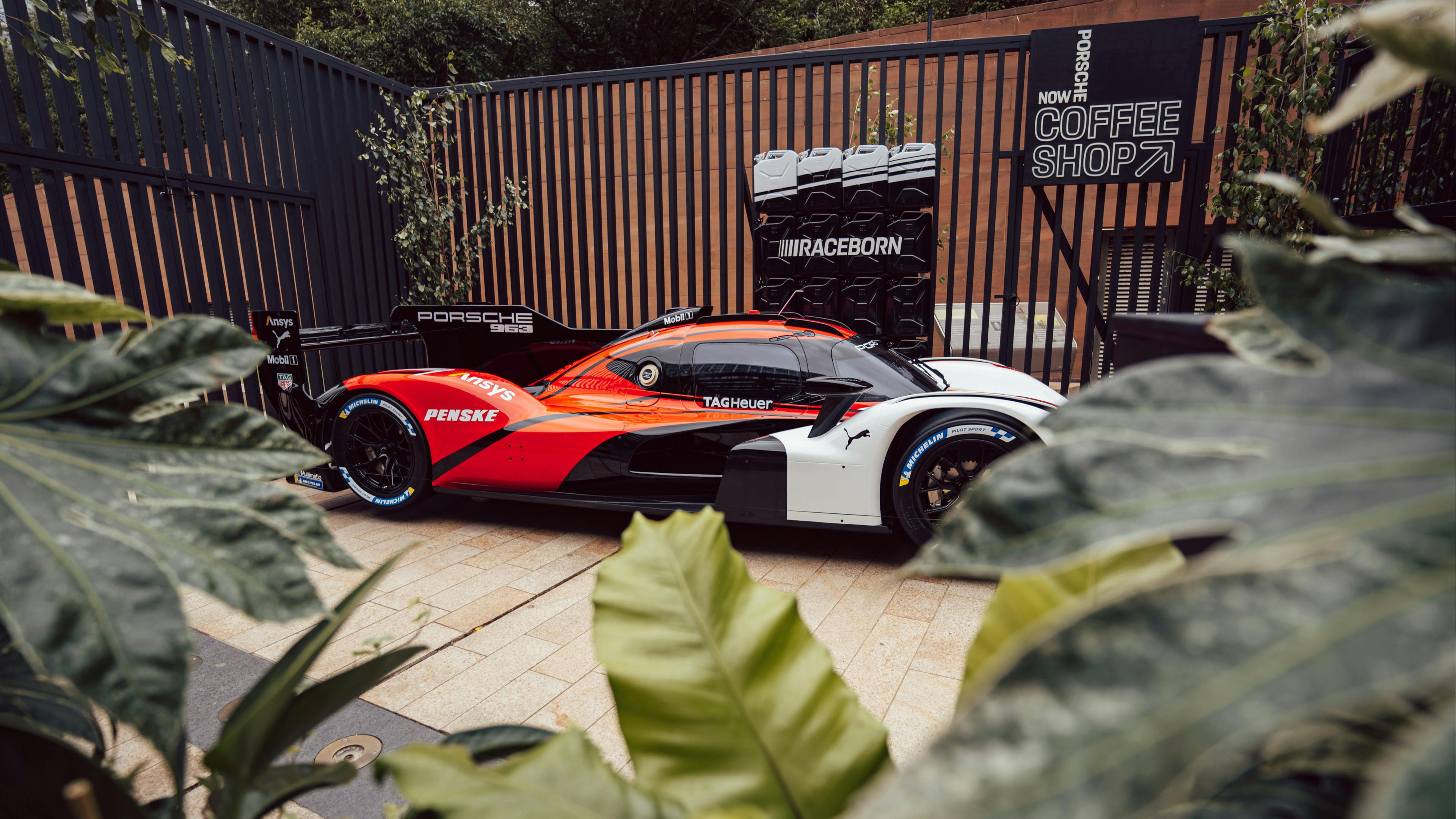 A high-performance Porsche 963 race car with red, black, and white livery parked in an outdoor courtyard. The shot is framed by green leaves in the foreground, with a “Porsche Now Coffee Shop” sign in the background.