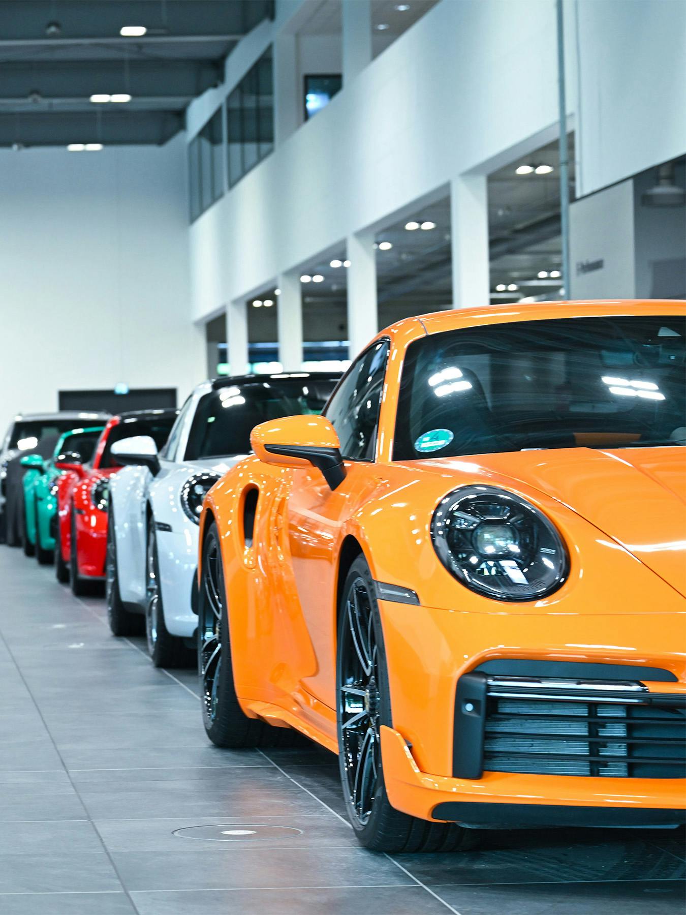 Orangener Porsche 911 im Vordergrund vor grauen, roten, türkisen und weißen Modellen im Showroom.