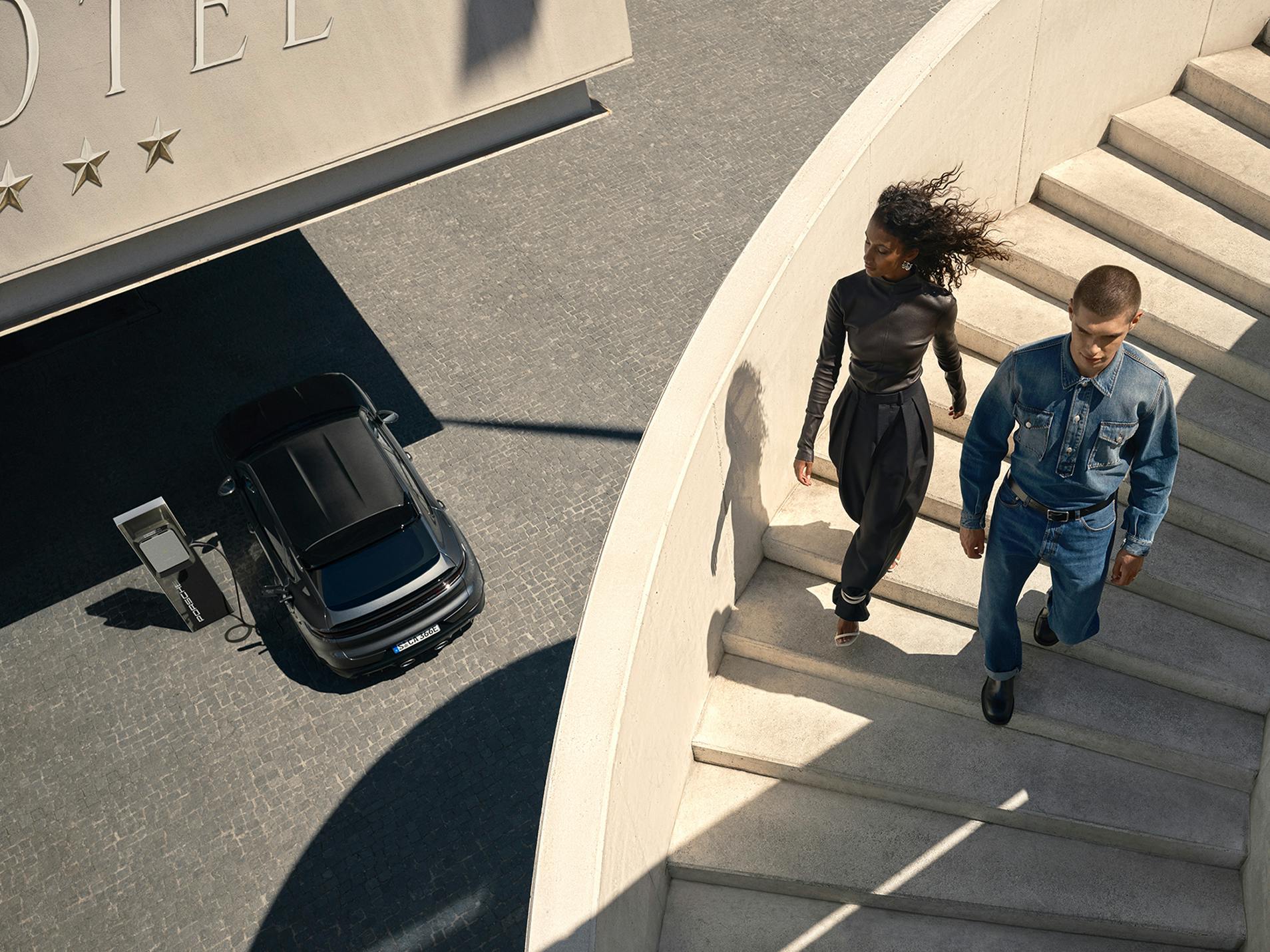 A woman and a man walking down a staircase. A Porsche Macan 4S is charging at a charging station below.