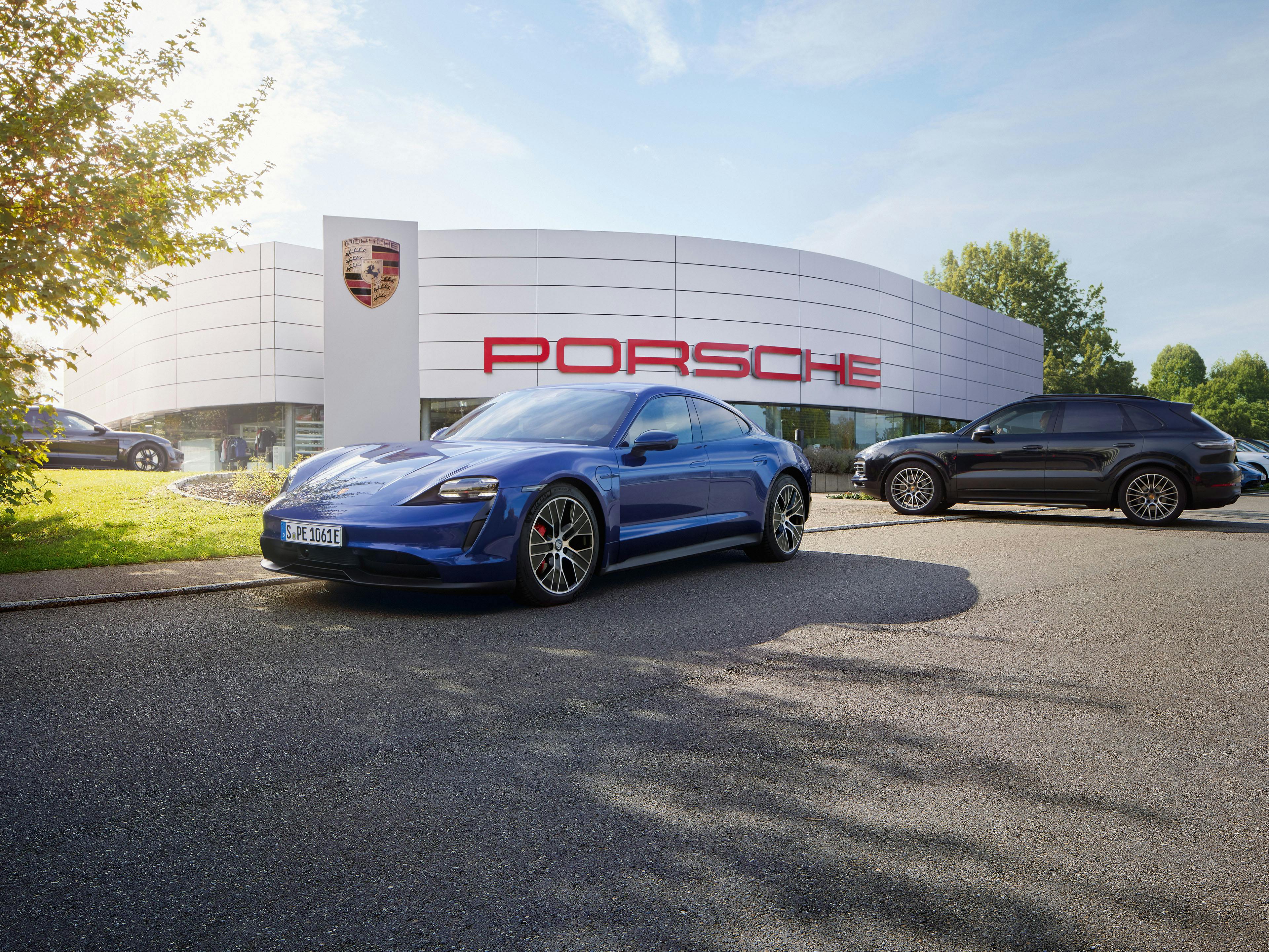Blauer Porsche Cayenne vor einem Porsche Zentrum mit rotem Porsche Schriftzug.