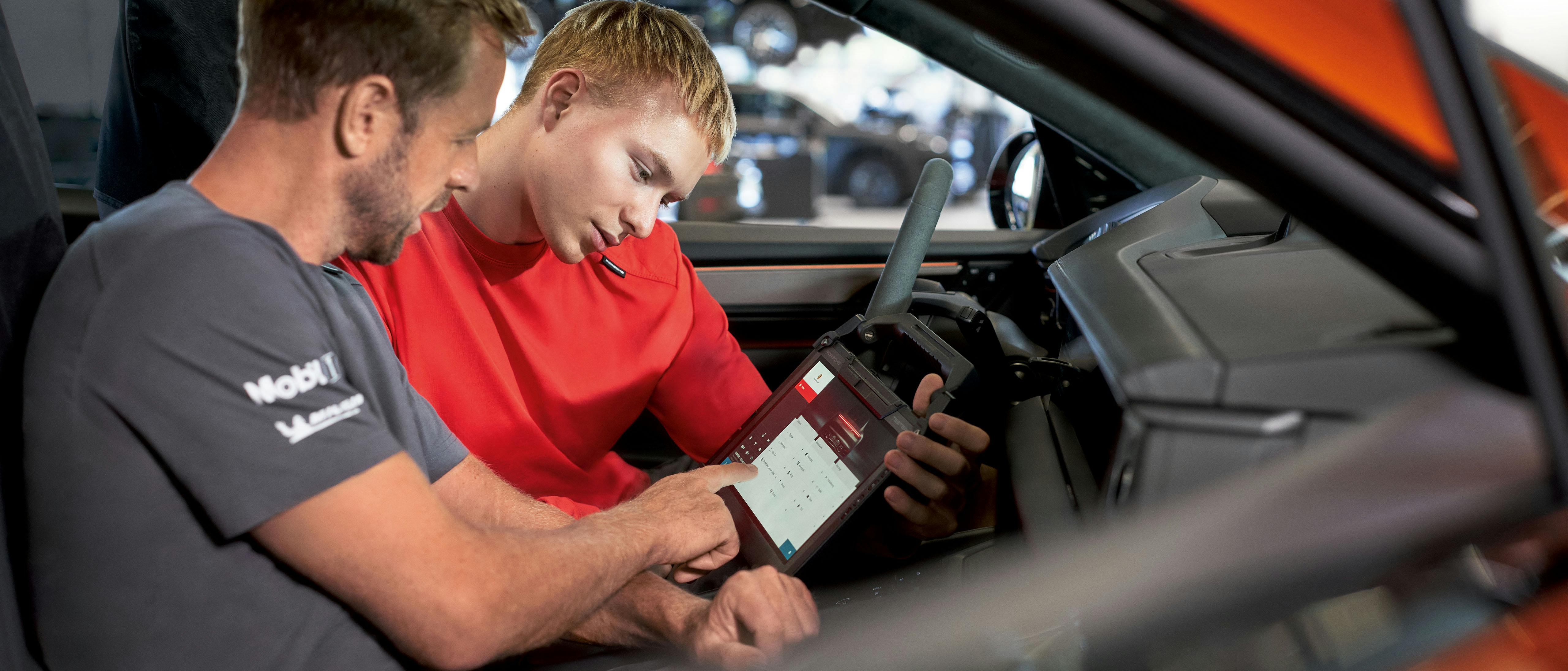 Twee monteurs zitten in een Porsche en kijken naar een tablet. De man in het grijze shirt wijst met zijn vinger naar het scherm terwijl de jongere man in het rode shirt aandachtig kijkt. Ze lijken samen een probleem te analyseren of een diagnose te stellen.