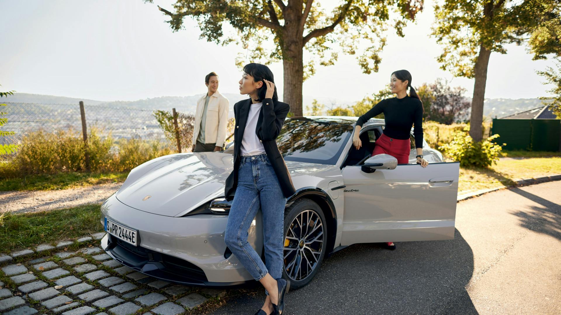 Two women and one man casually standing around a light grey Porsche Taycan on a sunny hillside road, with scenic views and trees in the background.