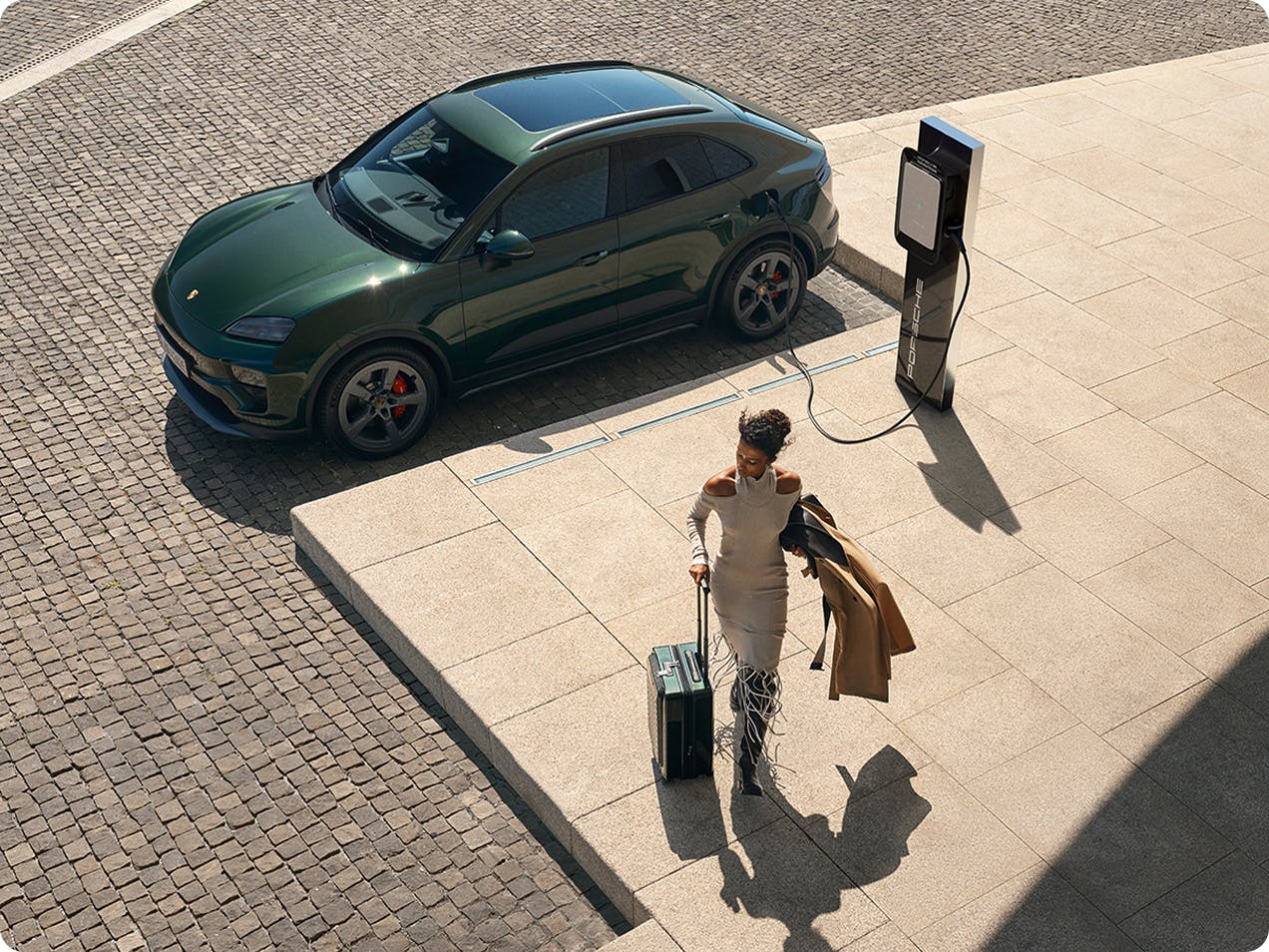Bird's-eye view of a Porsche Macan 4S in Oak Green Metallic being charged. A woman with a suitcase walks away from the car.