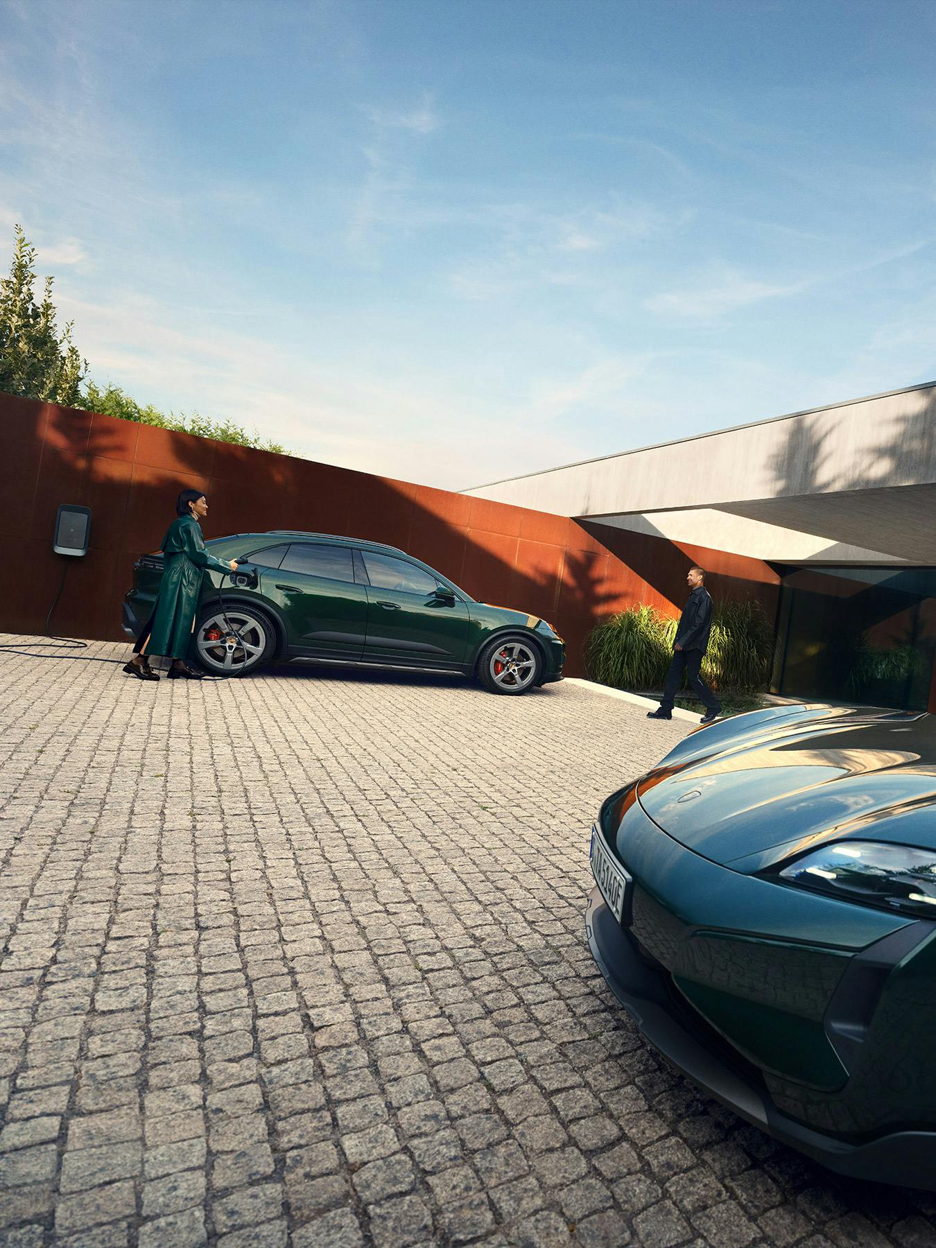 The Macan Electric and Taycan parked outside a house, with the Macan on charge in the background while the Taycan is parked in the foreground.