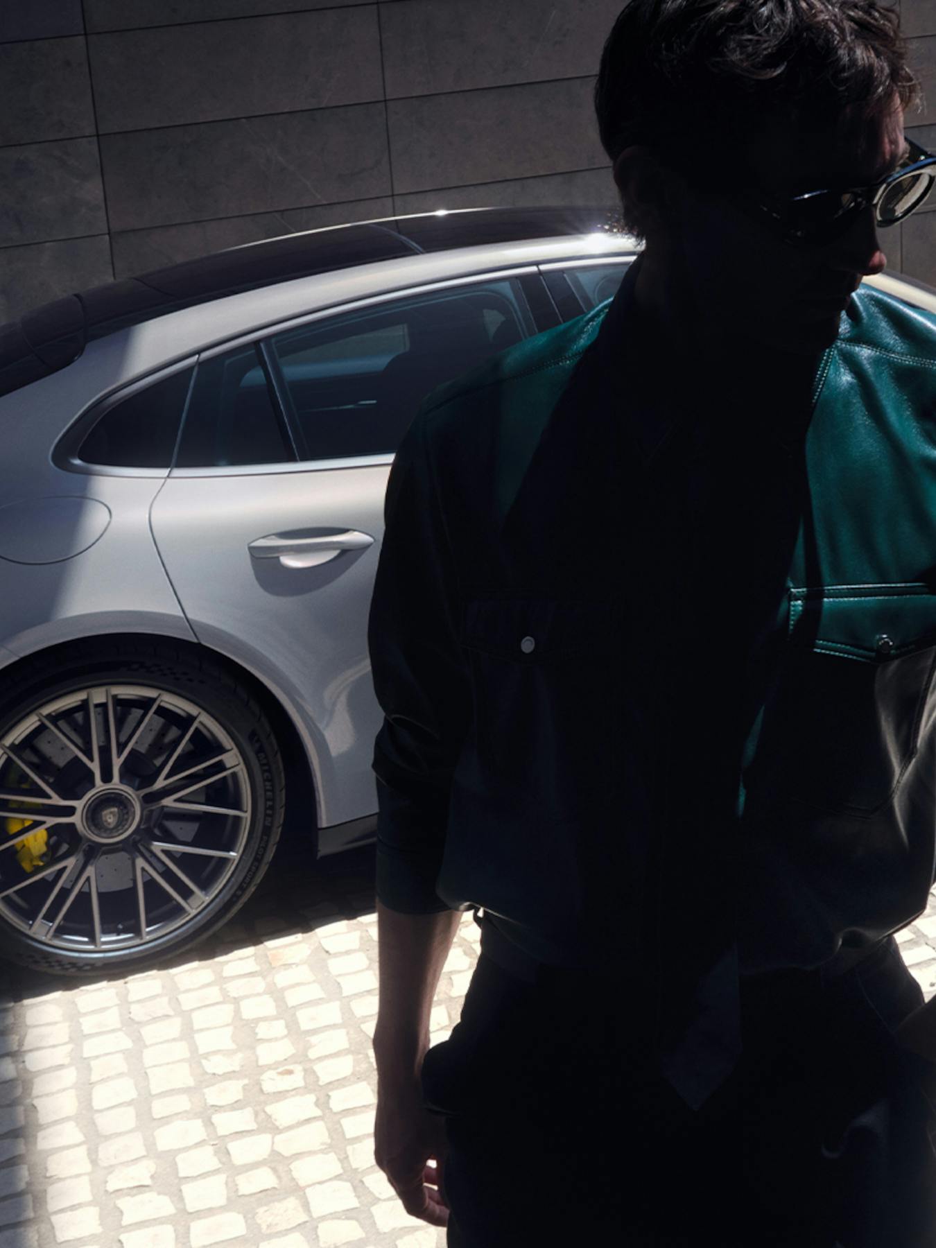 A high-angle shot showing a man standing in the foreground, particularly in shadow. In the background, a white Porsche Panamera with yellow brake calipers is parked on cobblestone surface next to a stone wall.​