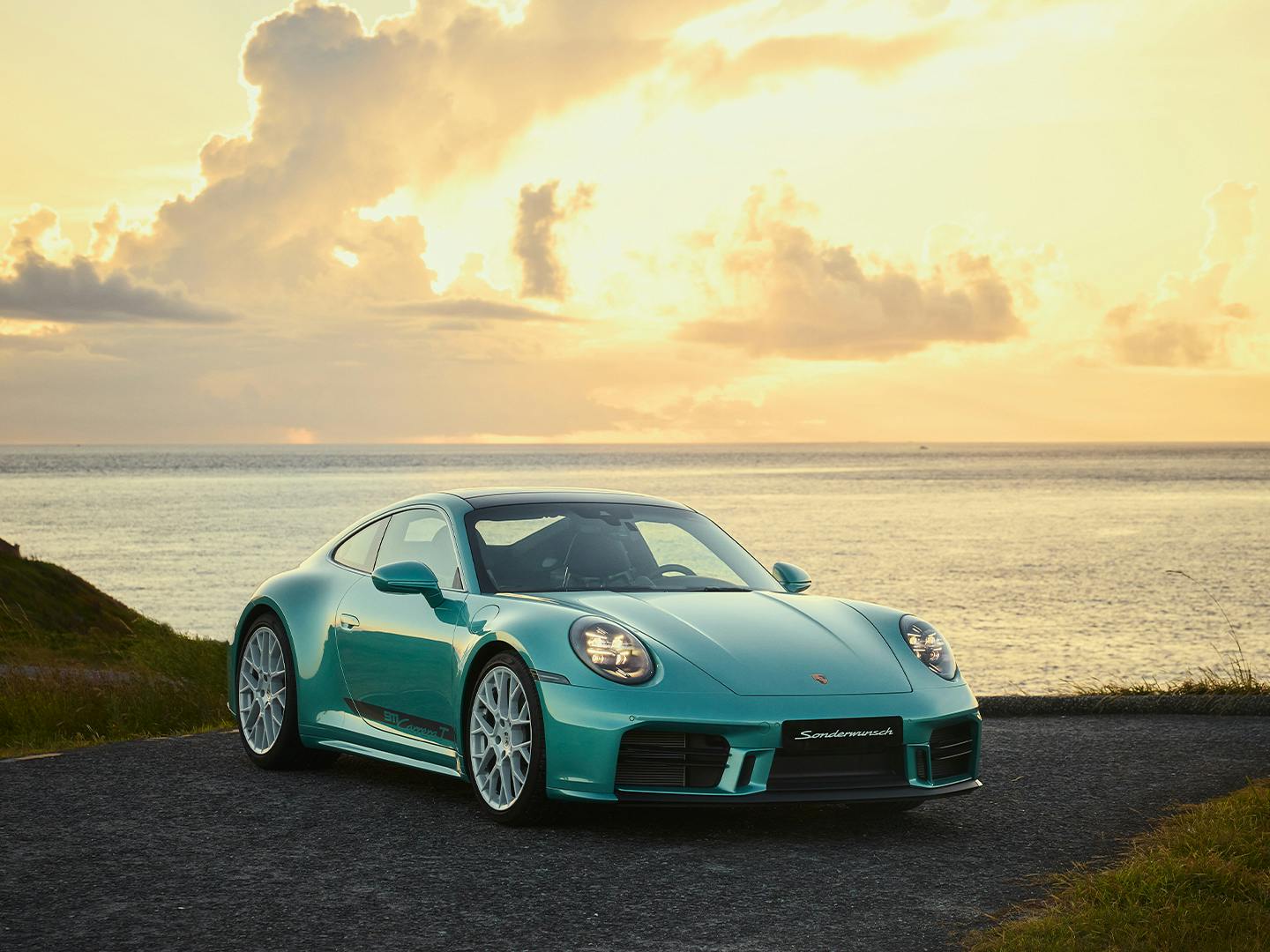 Detail of a turquoise Porsche 911 Carrera parked on a coastal road at sunset, front view with headlights visible.