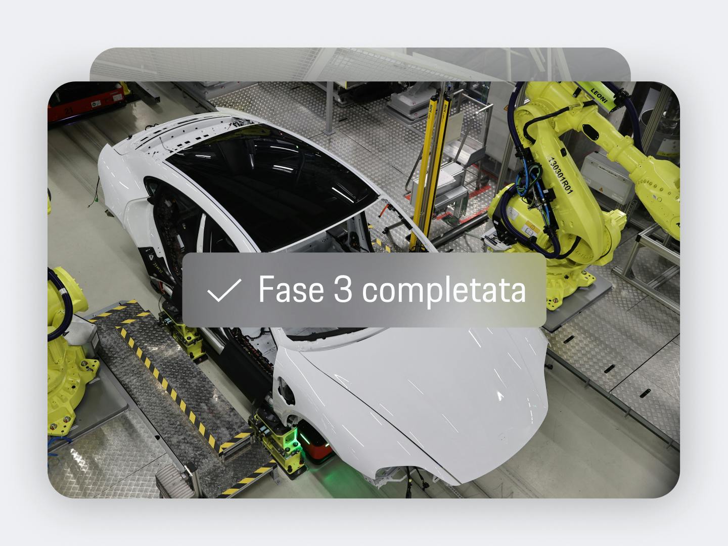 Vista dell'app che mostra la carrozzeria bianca di una Porsche sulla linea di assemblaggio con bracci robotici gialli e testo sovrapposto “Fase 3 completata”.