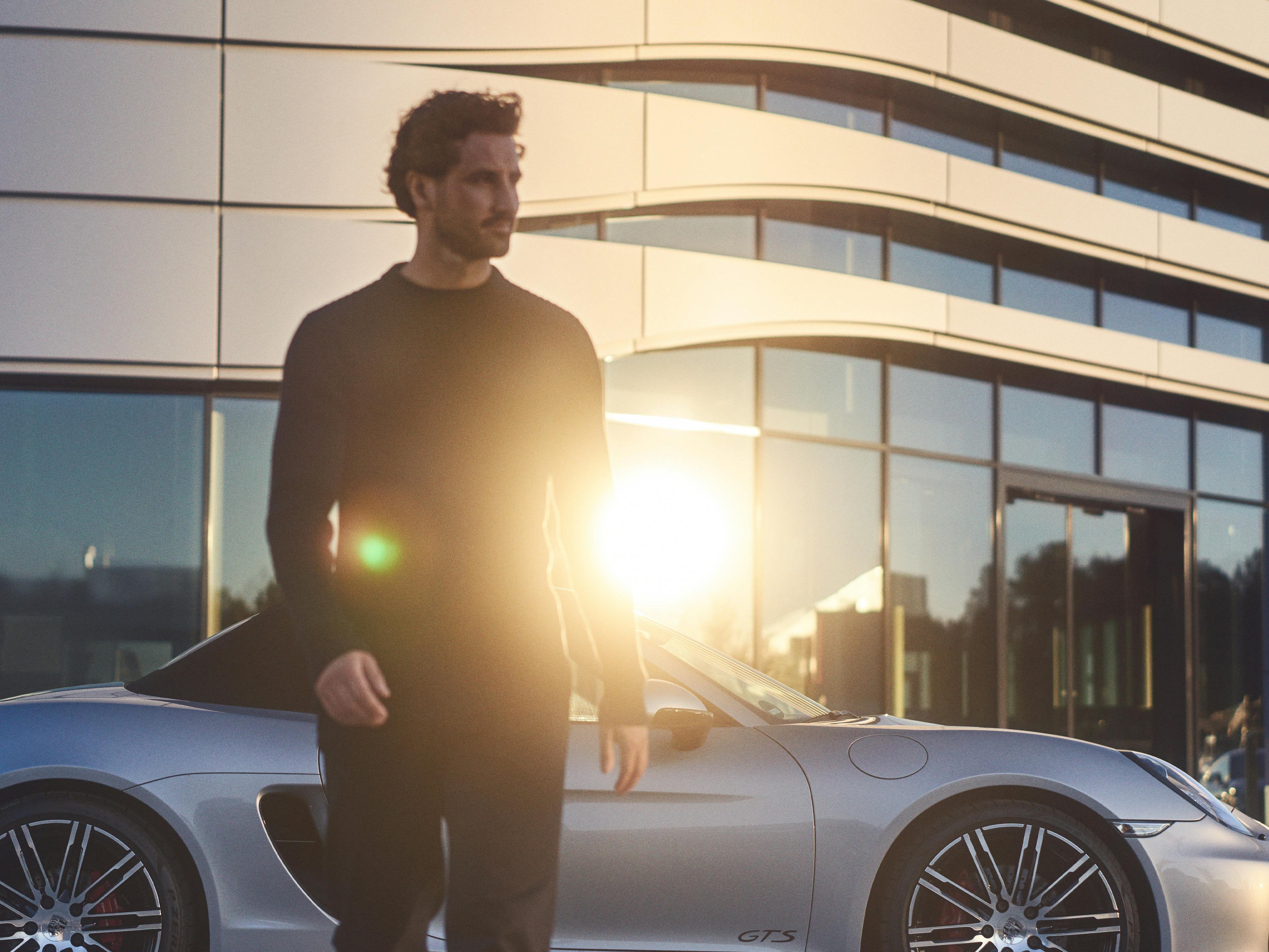 A man in a black outfit stands near a silver Porsche sports car with modern architecture and sunlight in the background.