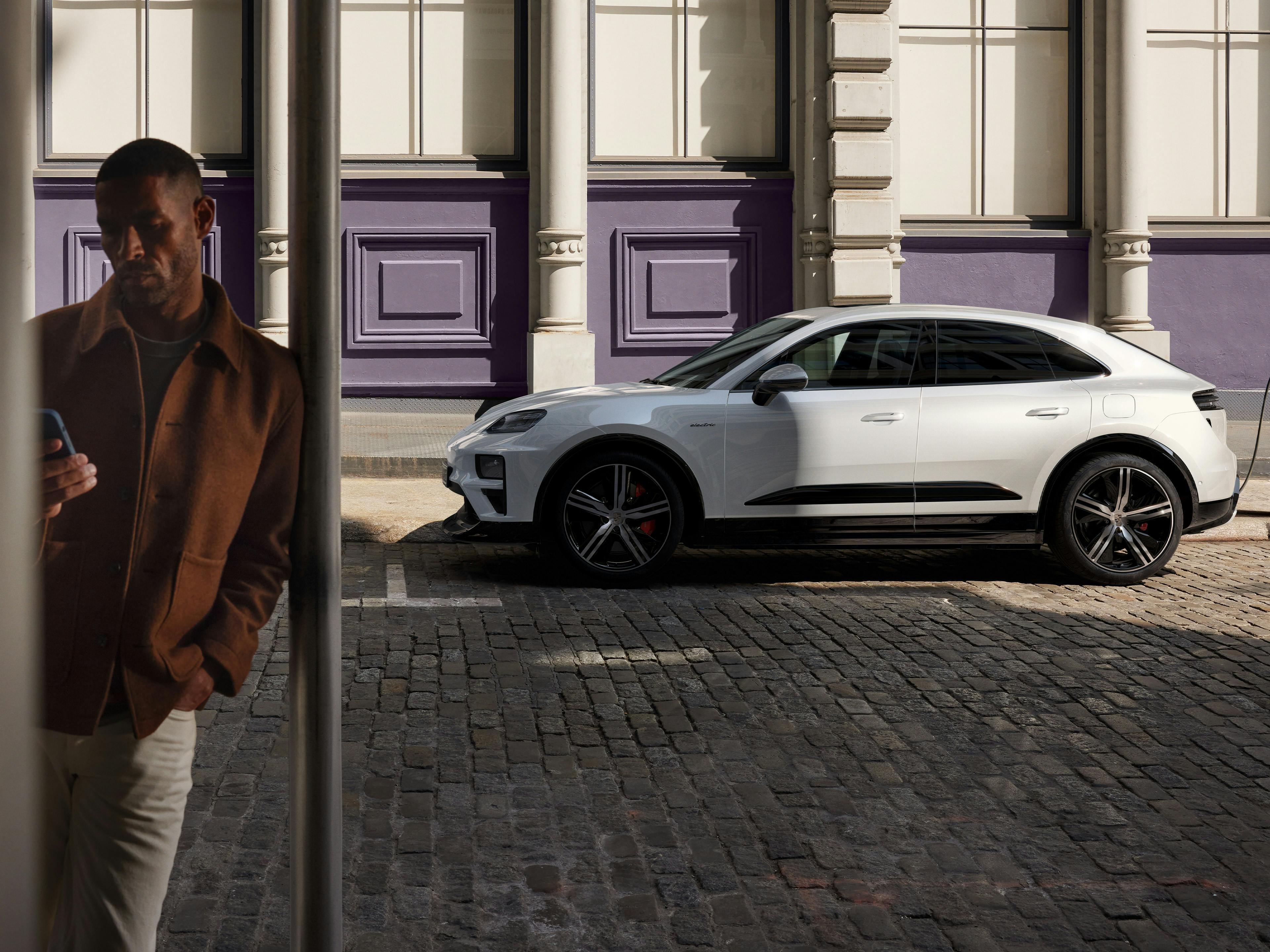 Ein Mann steht gelehnt an einer Stange und schaut auf sein Hand. Im Hintergund parkt ein Porsche Macan.