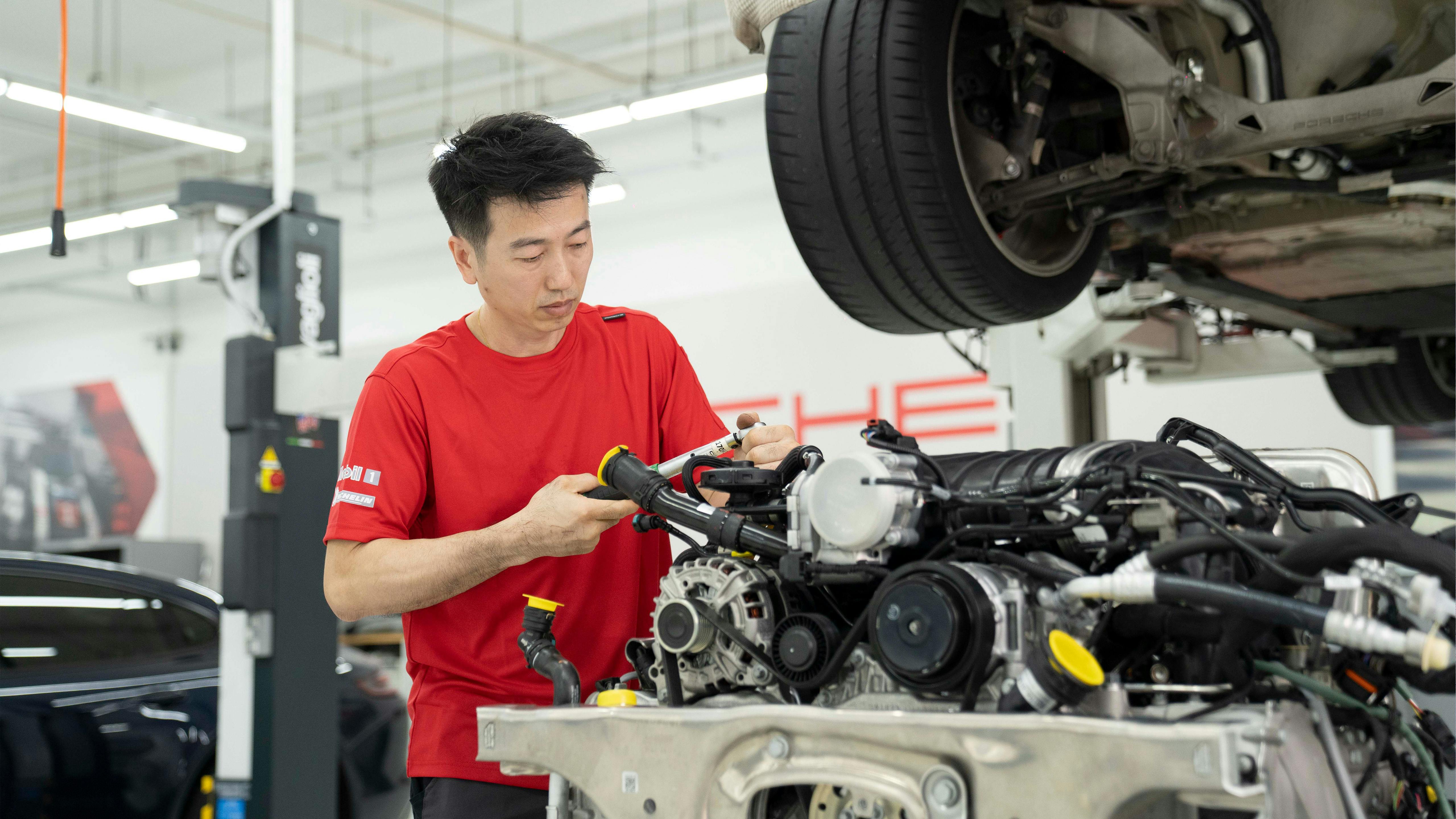Porsche mechanic working intently on a removed engine in a bright workshop with a car lift.