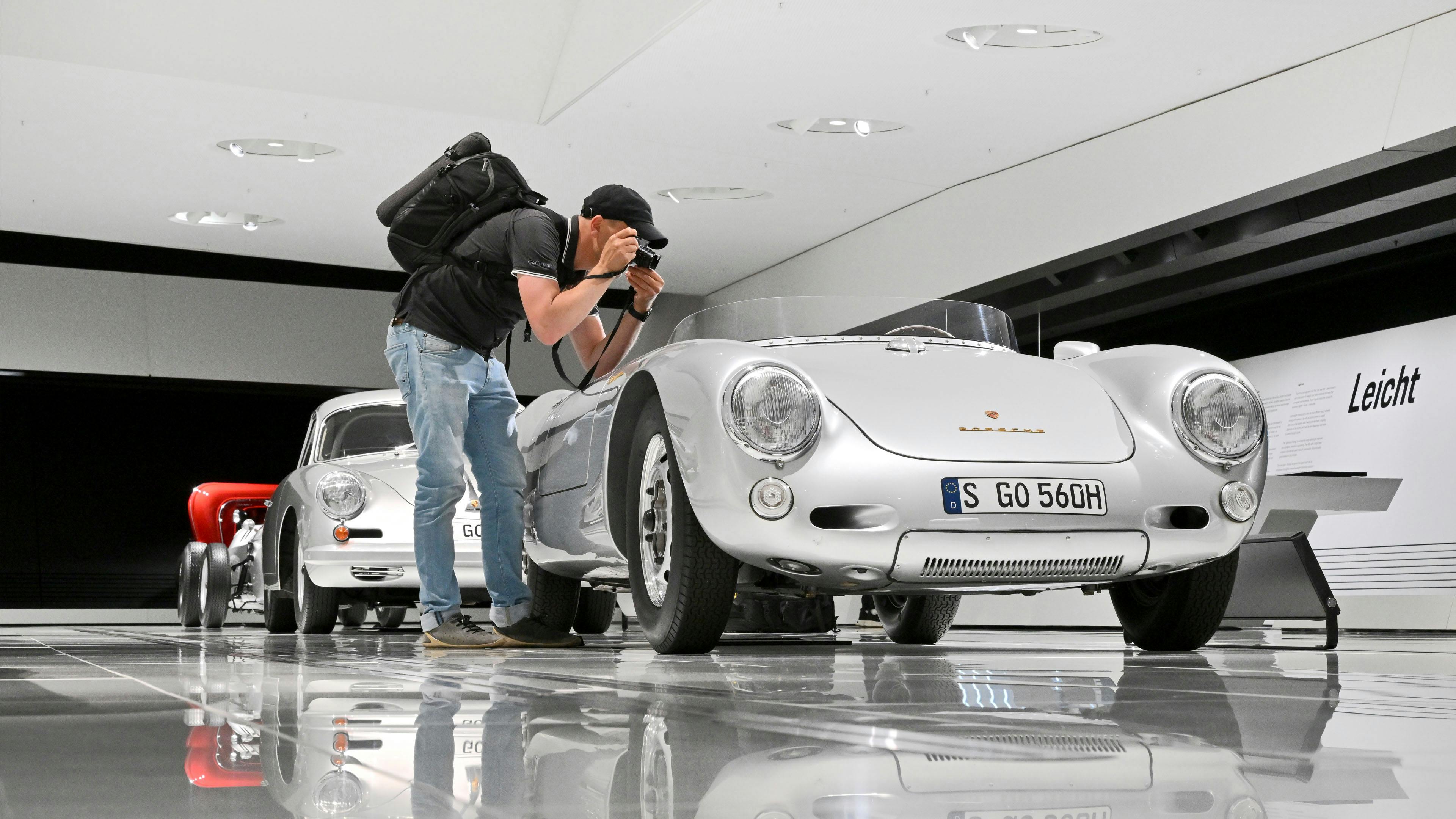 Ein Besucher mit Kamera und Rucksack fotografiert einen historischen silbernen Porsche‑Sportwagen im Ausstellungsraum des Museums.