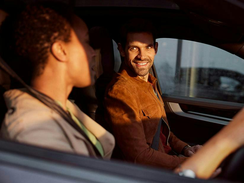A man in a brown suede jacket sits in the passenger seat of a car, smiling as he looks toward the driver. The scene is bathed in the warm, golden light of a sunset or sunrise, visible through the side window.