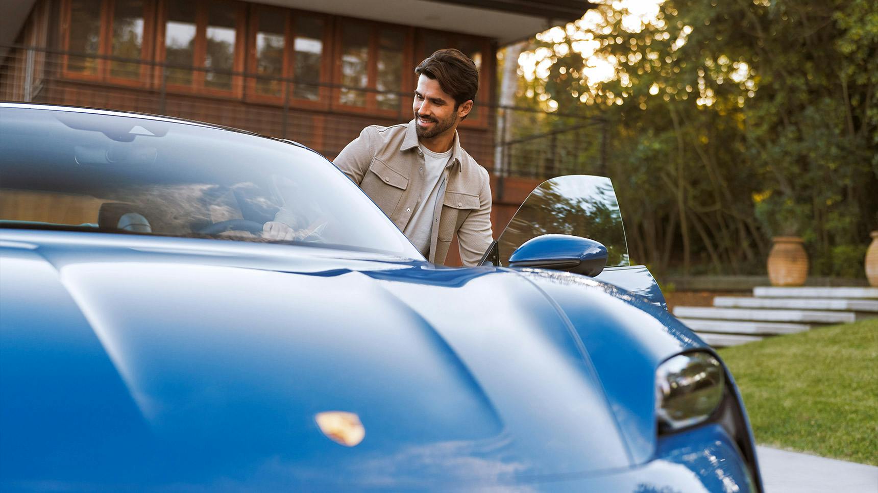 A vibrant blue Porsche sports car parked near a modern house, with a man opening the driver's door.