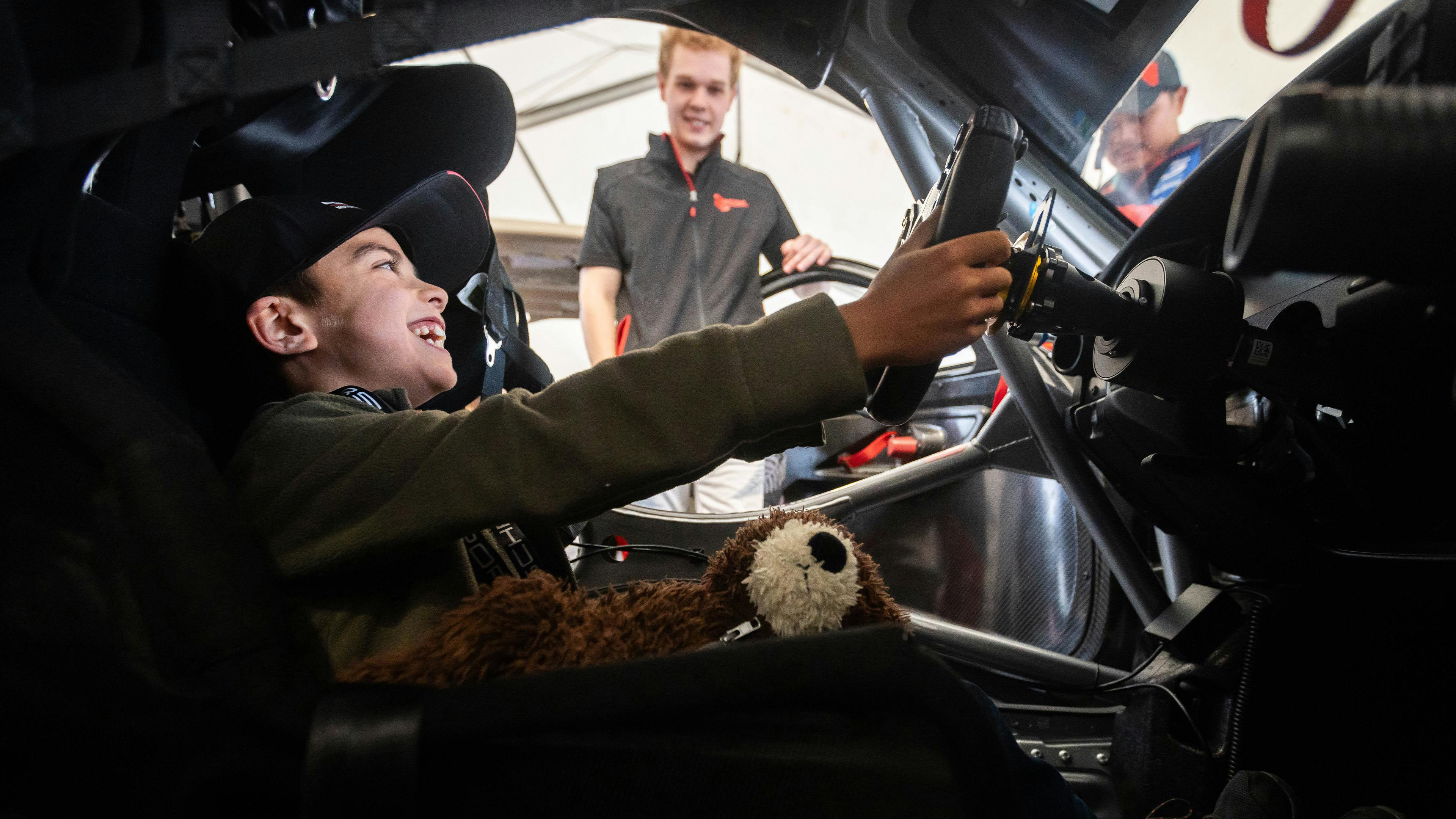 A smiling child with short hair sits in the driver's seat of a Porsche. Another individual is partially visible in the background.
