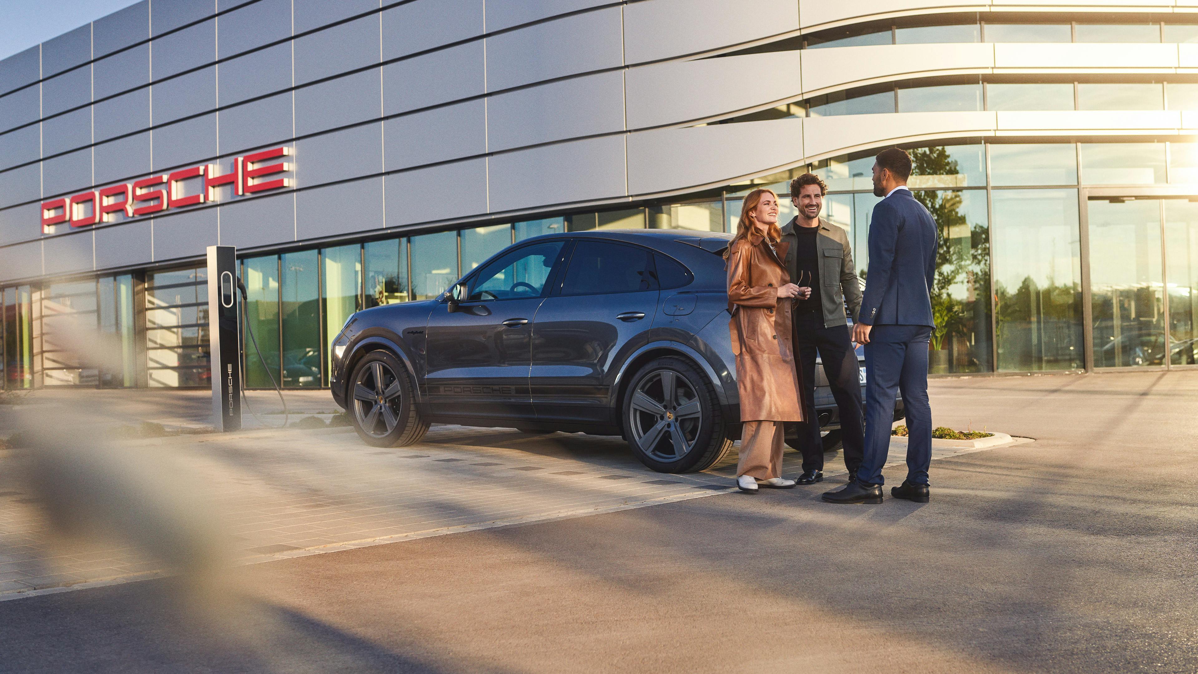 A couple and a Porsche consultant have a conversation outside a Porsche Centre. A Porsche employee and a couple are having a consultation. A Porsche can be seen in the background. In the foreground is a Mobil 1 oil bottle. A Porsche 911 can be seen in the background.