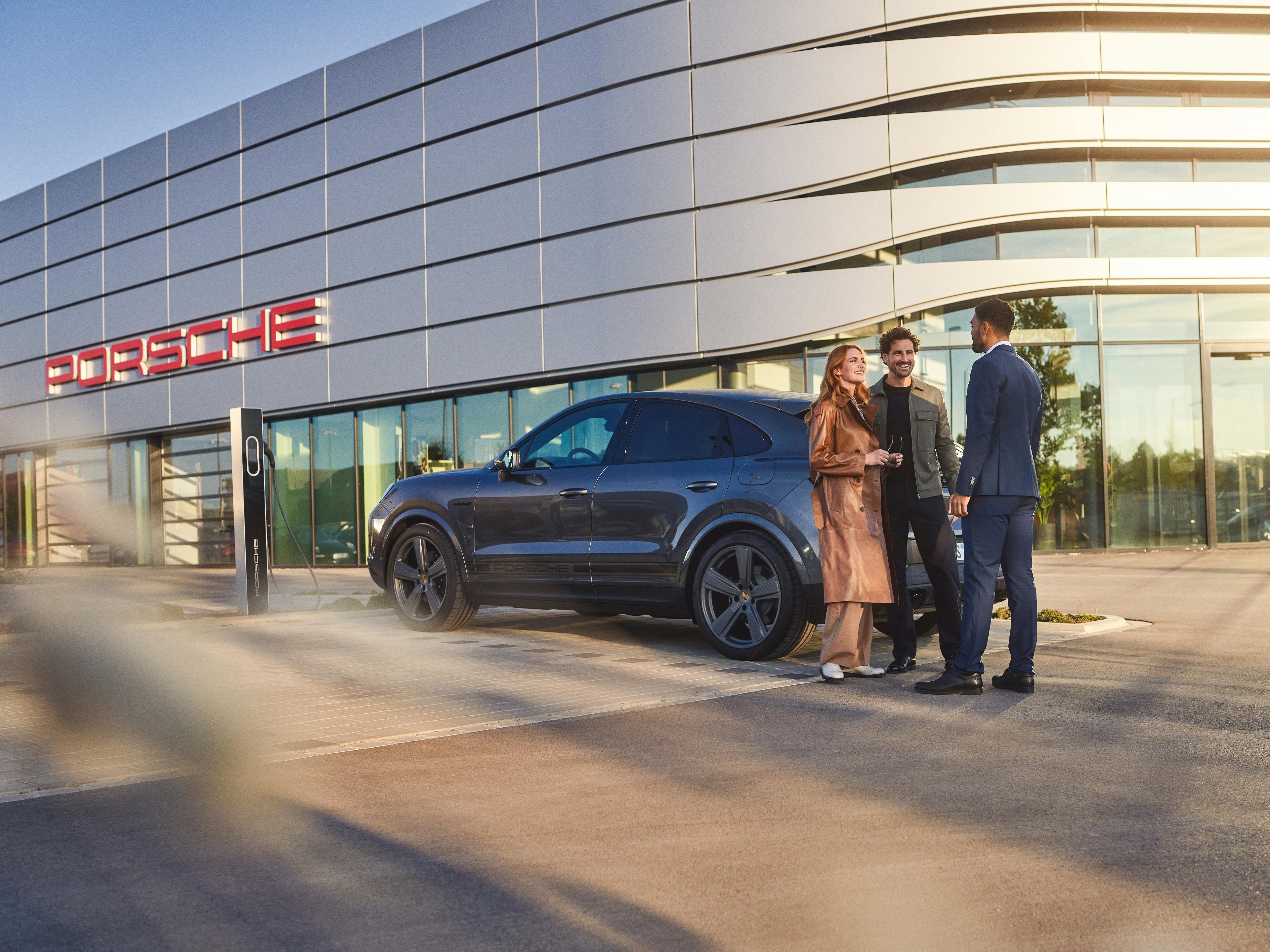 Ein Porsche Mitarbeiter und ein Paar stehen vor einem Porsche-Showroom neben einem Porsche SUV