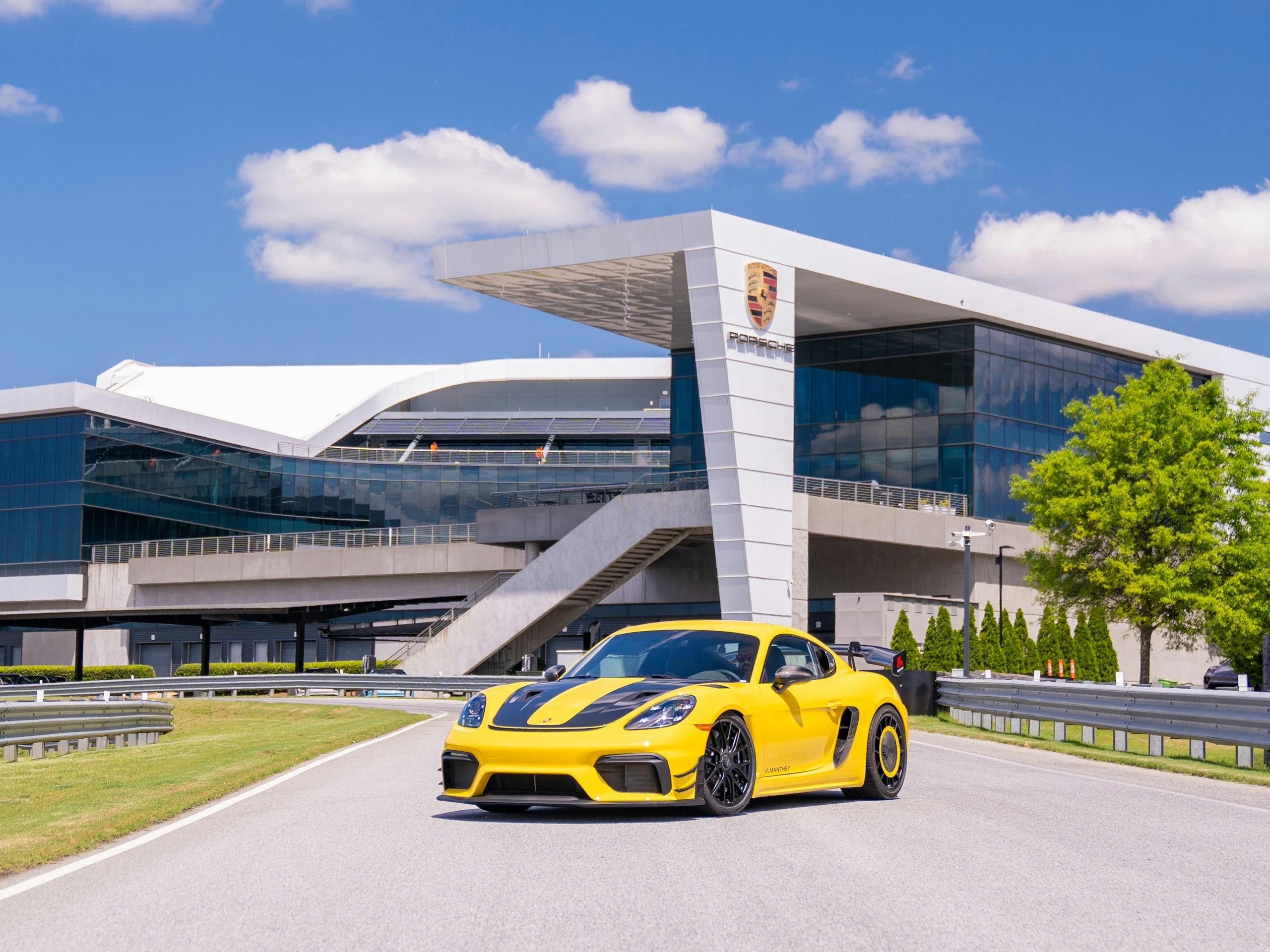 A yellow Cayman GT4 RS with Manthey Package sits on a road in front of a Porsche building