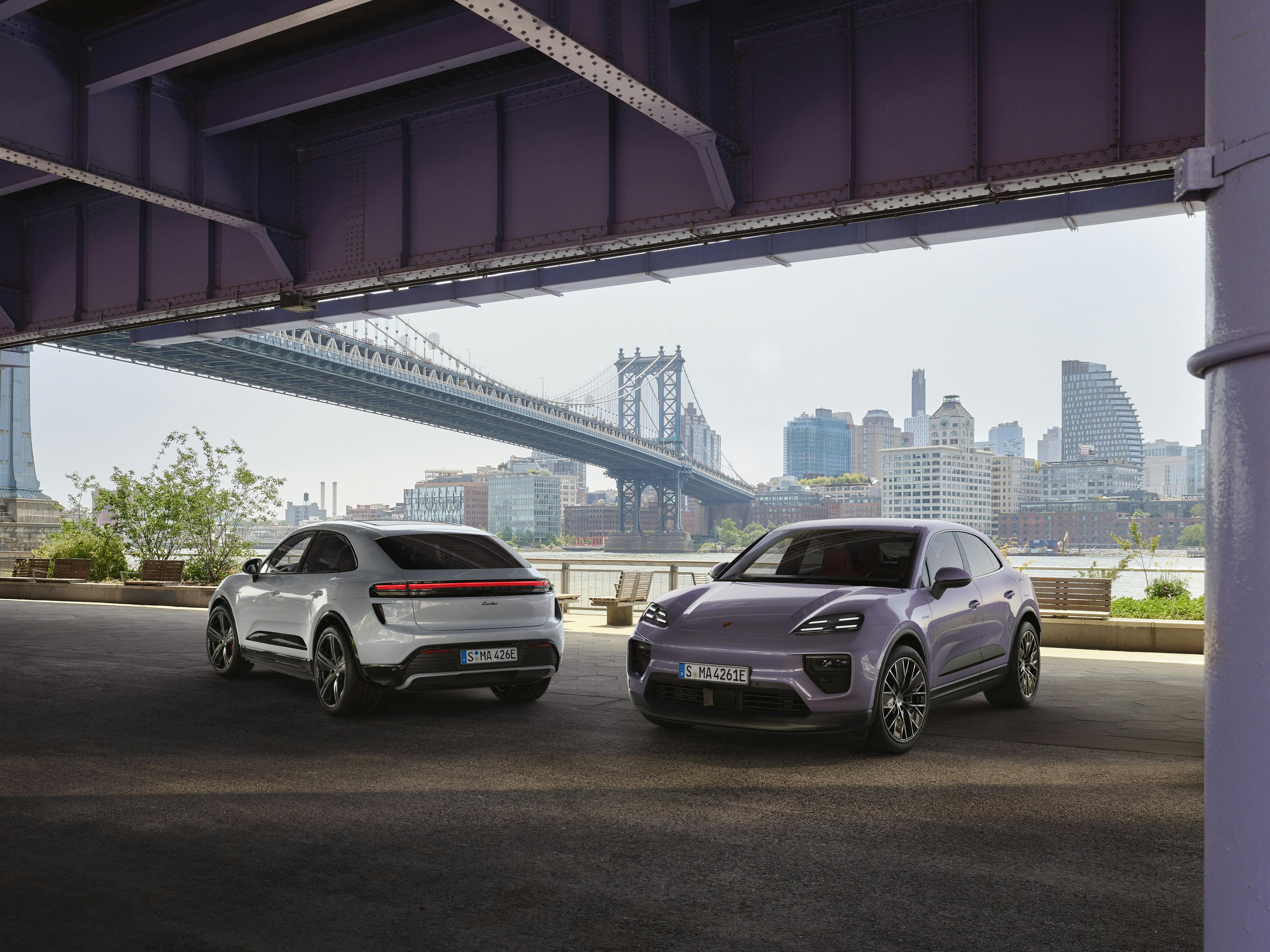 Zwei Porsche Macan Elektro, einer weiß und der andere lila, sind unter einer eisernen Brücke geparkt, mit einer Stadtansicht im Hintergrund.