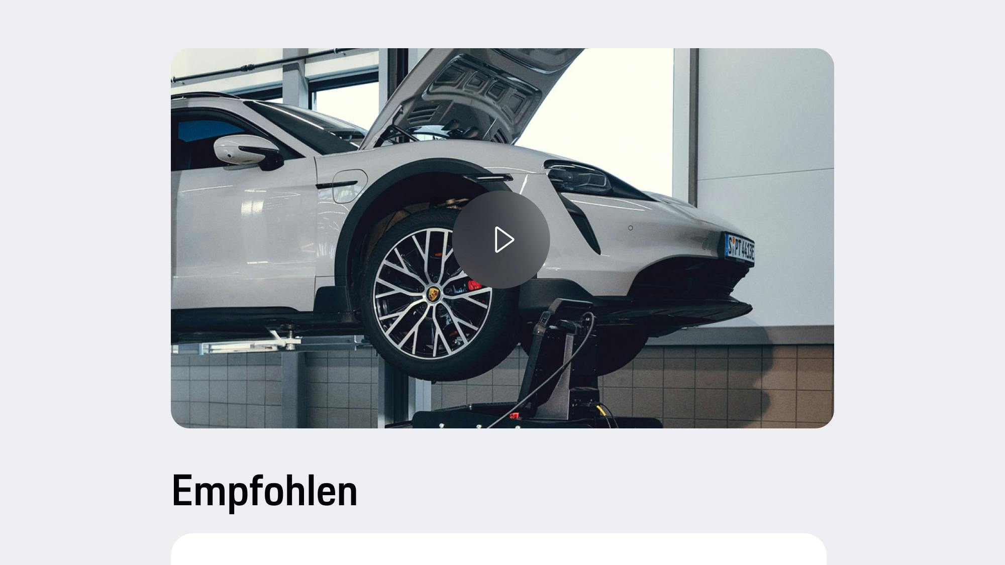 App-Ansicht mit hellgrauem Porsche auf Hebebühne mit geöffneter Motorhaube, Video-Wiedergabetaste und darunter angezeigter Empfehlungstext.
