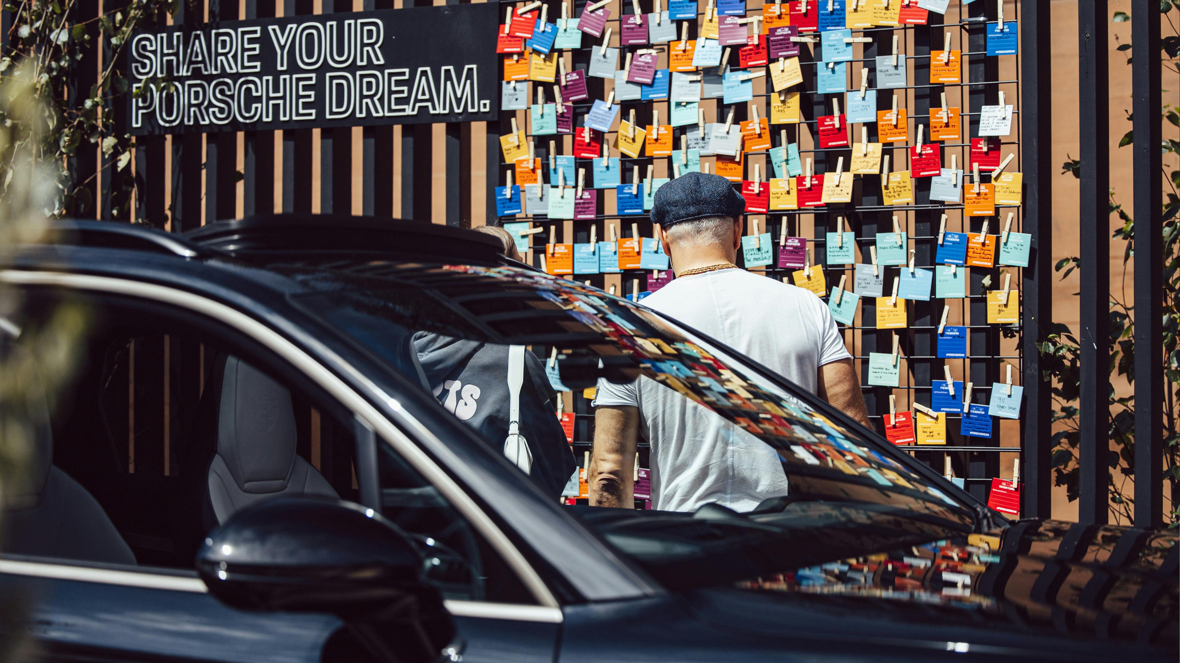 A person stands in front of a black slatted wall labelled “Share Your Dream Porsche”, which is covered in colourful small notes clipped to a wire grid. A dark Porsche is partially visible in the foreground.