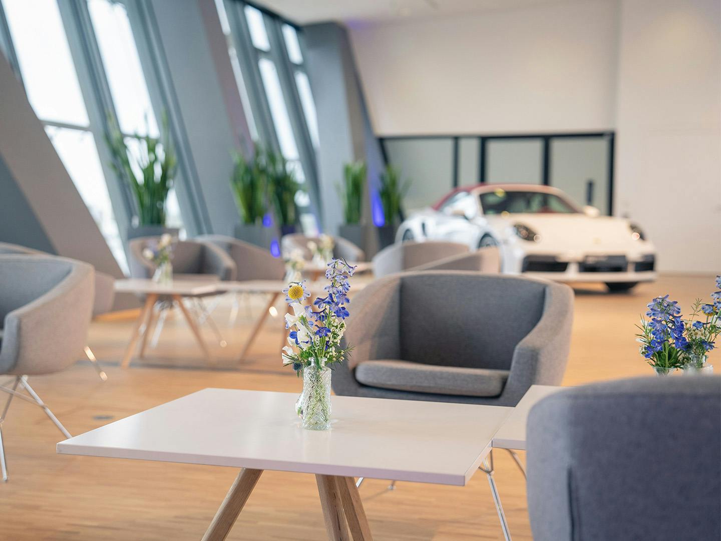 Im Vordergrund steht eine Blumenvase auf einem kleinen Tisch, der von Loungesesseln umgeben ist. Im Hintergrund erkennt man die Silhouette eines Porsche 911.