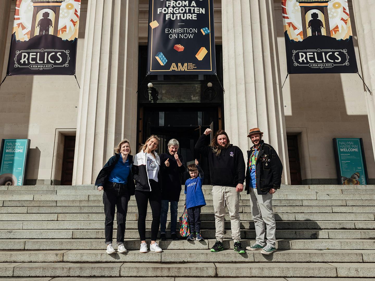 A group of six people of various ages standing on the steps of a grand building with large columns.