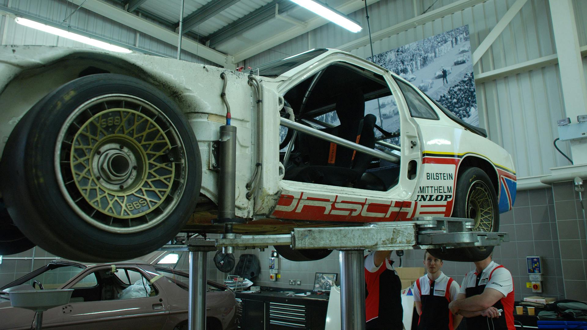 Multiple employees working on the bottom side of the Porsche 924 GTP which is standing on a lifting platform.