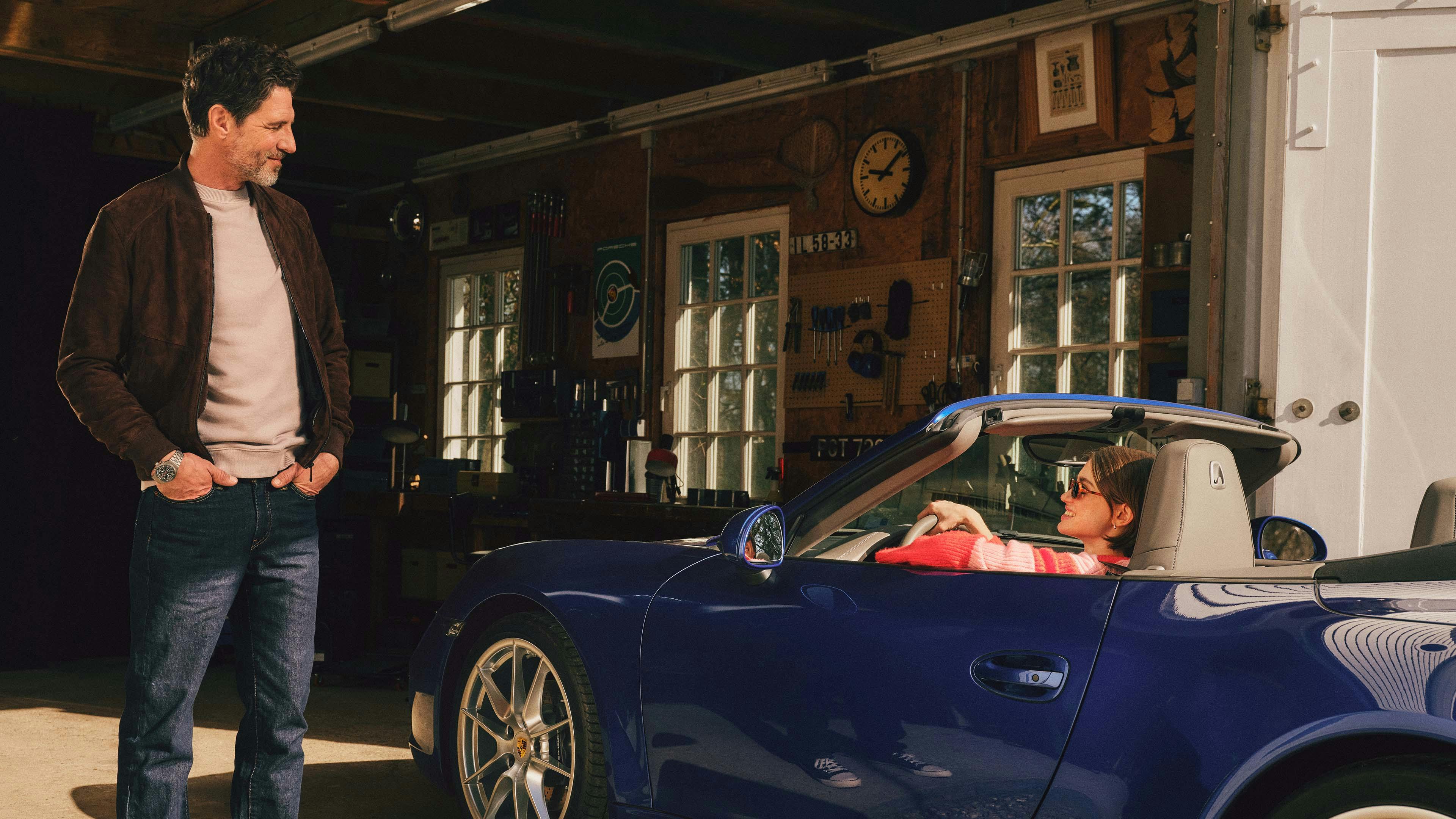 A blue Porsche 911 Cabriolet drives into a garage; a person is standing at the entrance looking at the vehicle