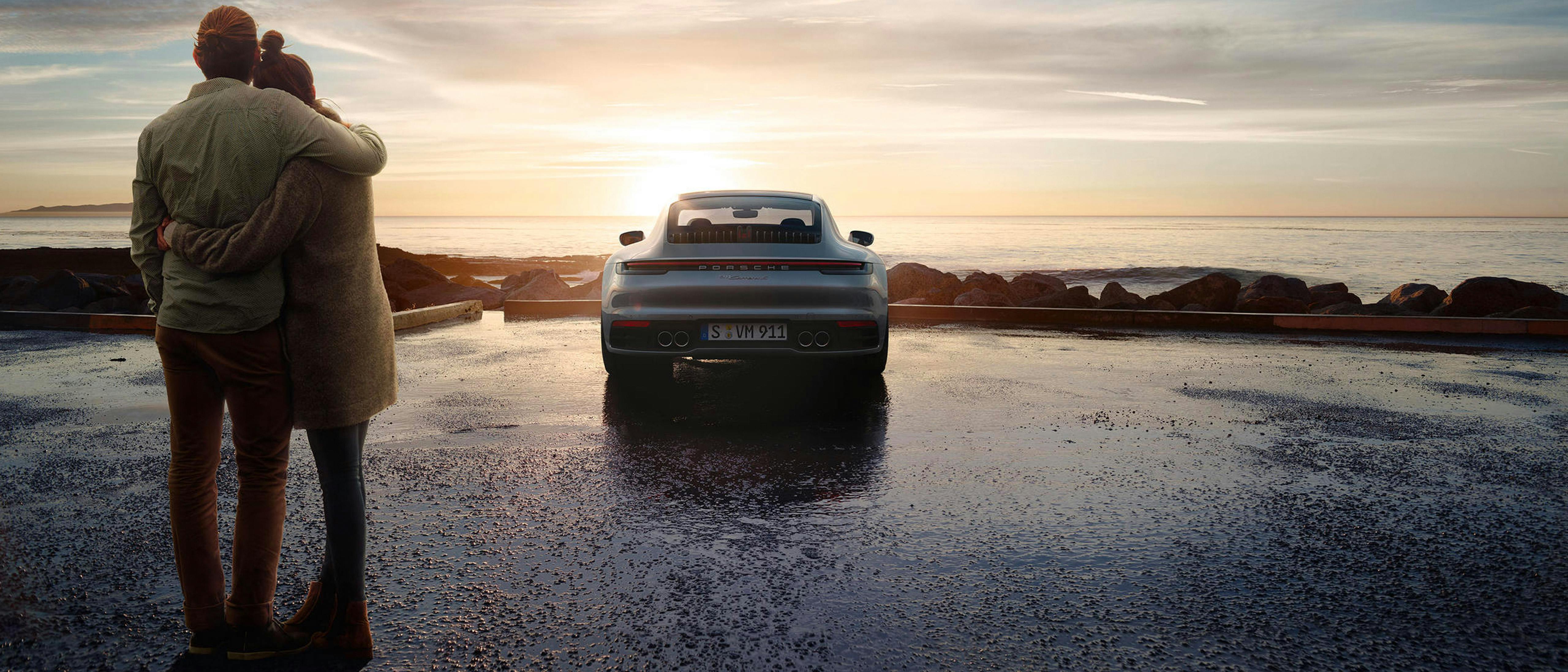Una coppia si abbraccia comodamente al tramonto in riva al mare davanti a una Porsche 911, mentre sullo sfondo si vede il mare calmo.