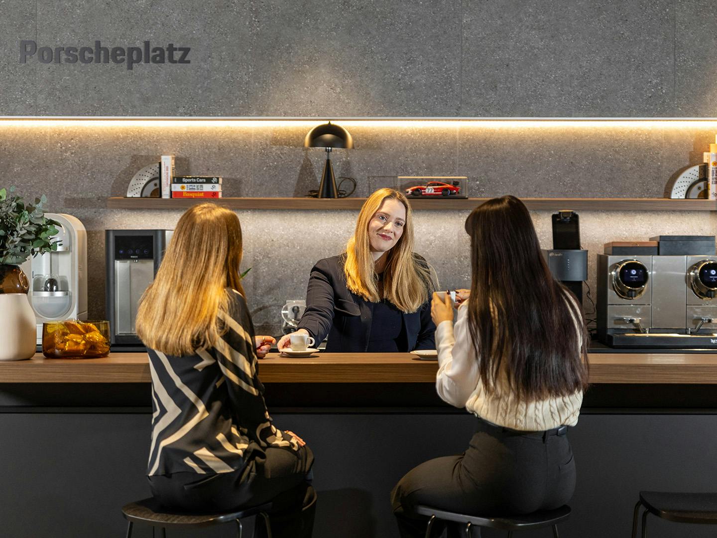 Une employée de Porsche, debout derrière le comptoir d'un café, sert du café à deux clientes assises devant le comptoir sur des tabourets de bar.