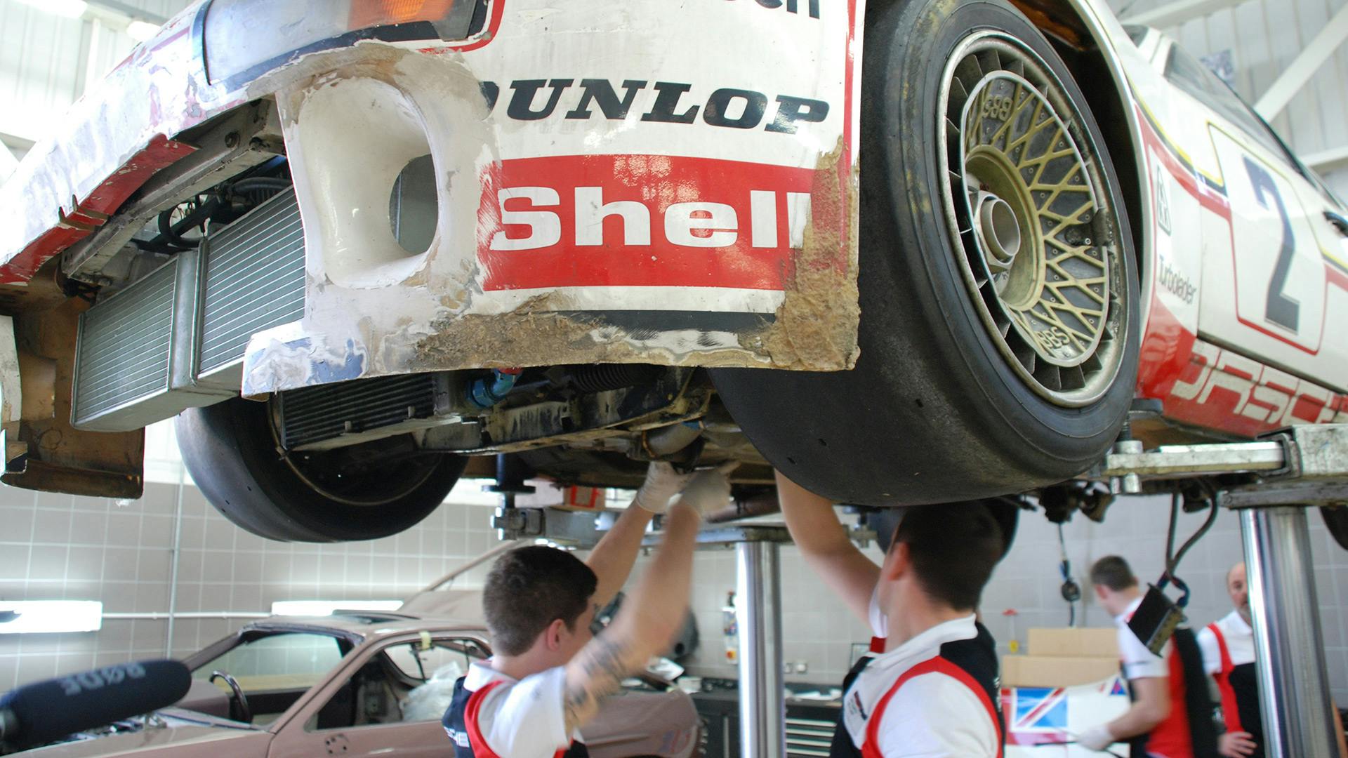 Multiple employees working on the bottom side of the Porsche 924 GTP which is standing on a lifting platform.