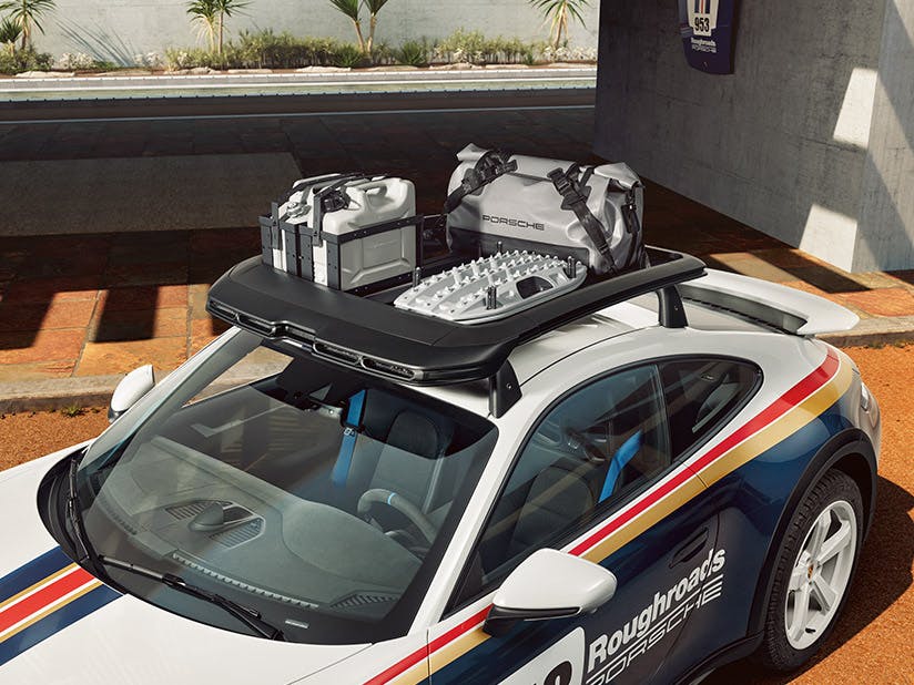 Un panier de toit est installé sur une Porsche 911 Dakar garée devant un batiment. Il contient des bidons d'huile, une plaque de déselisement et un sac Lifestyle.
